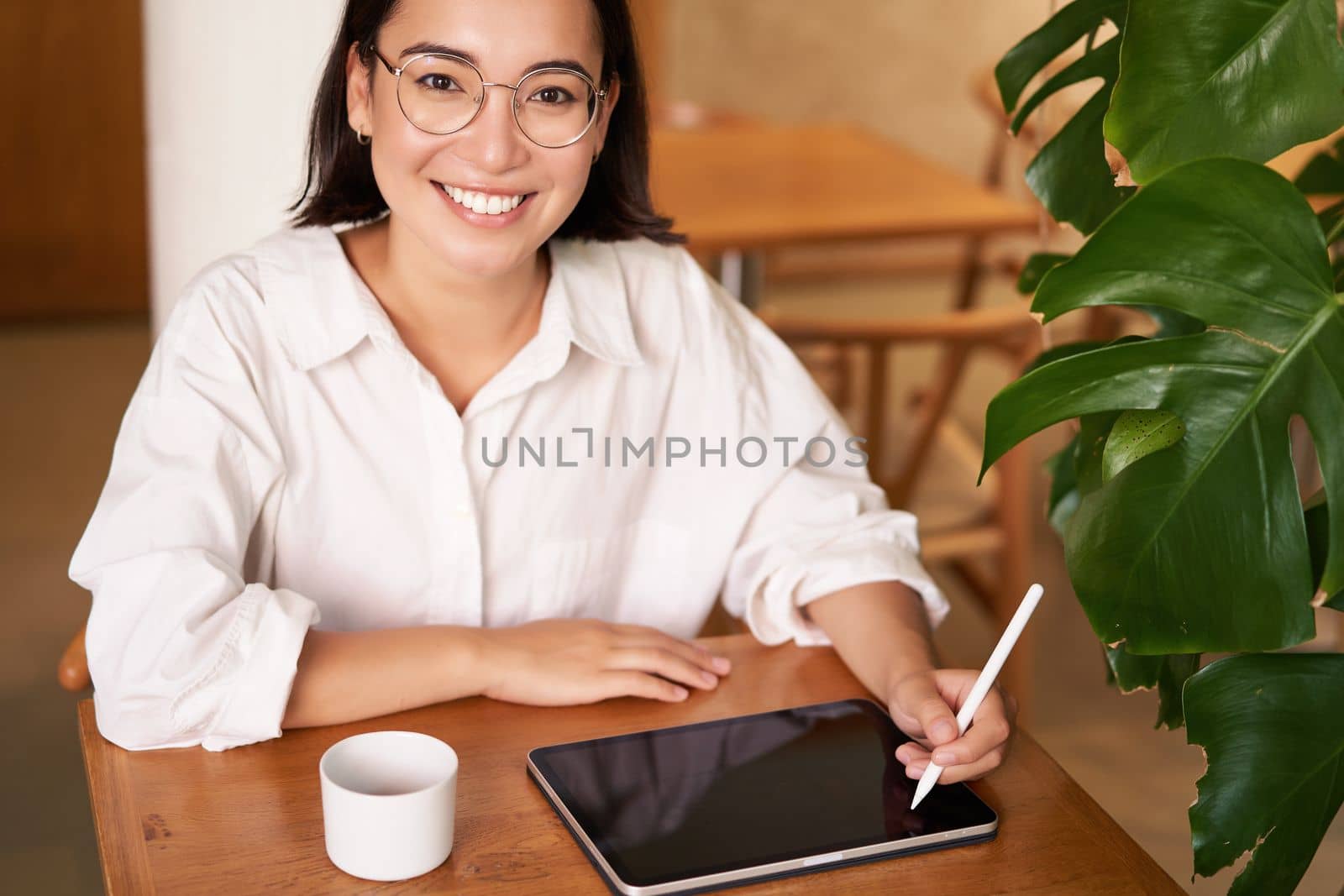 Smiling young female creator, creative girl drawing on digital tablet with graphic pen, drinking coffee in cafe.