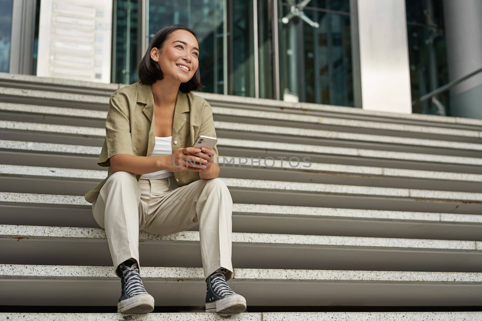 Portrait of happy smiling asian woman, sitting outdoors near building, using smartphone. Technology concept.