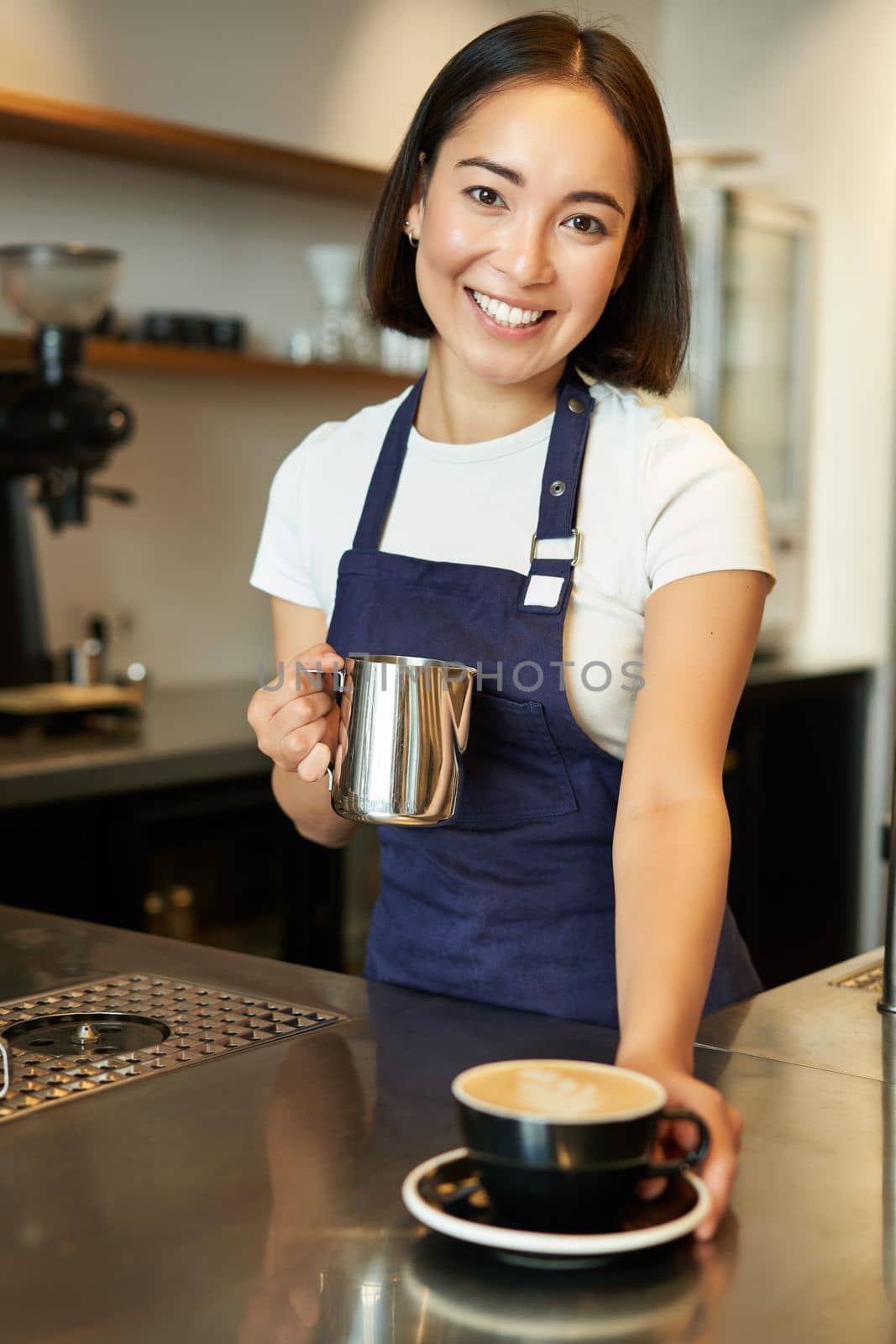 Vertical shot of smiling asian girl in cafe, giving out cappuccino, made order, prepared coffee for you, standing behind counter in blue apron.