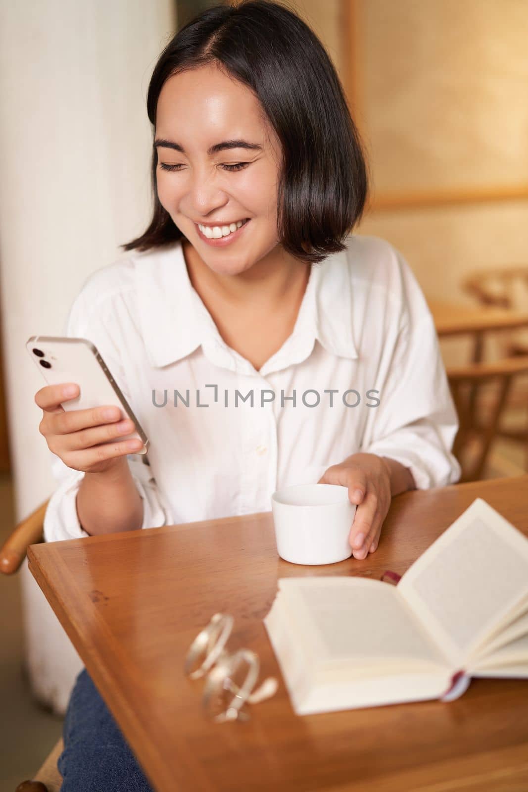 Happy asian woman sits in cafe with cup of coffee and book, answer video call on smartphone, laughing and talking via camera on mobile phone.