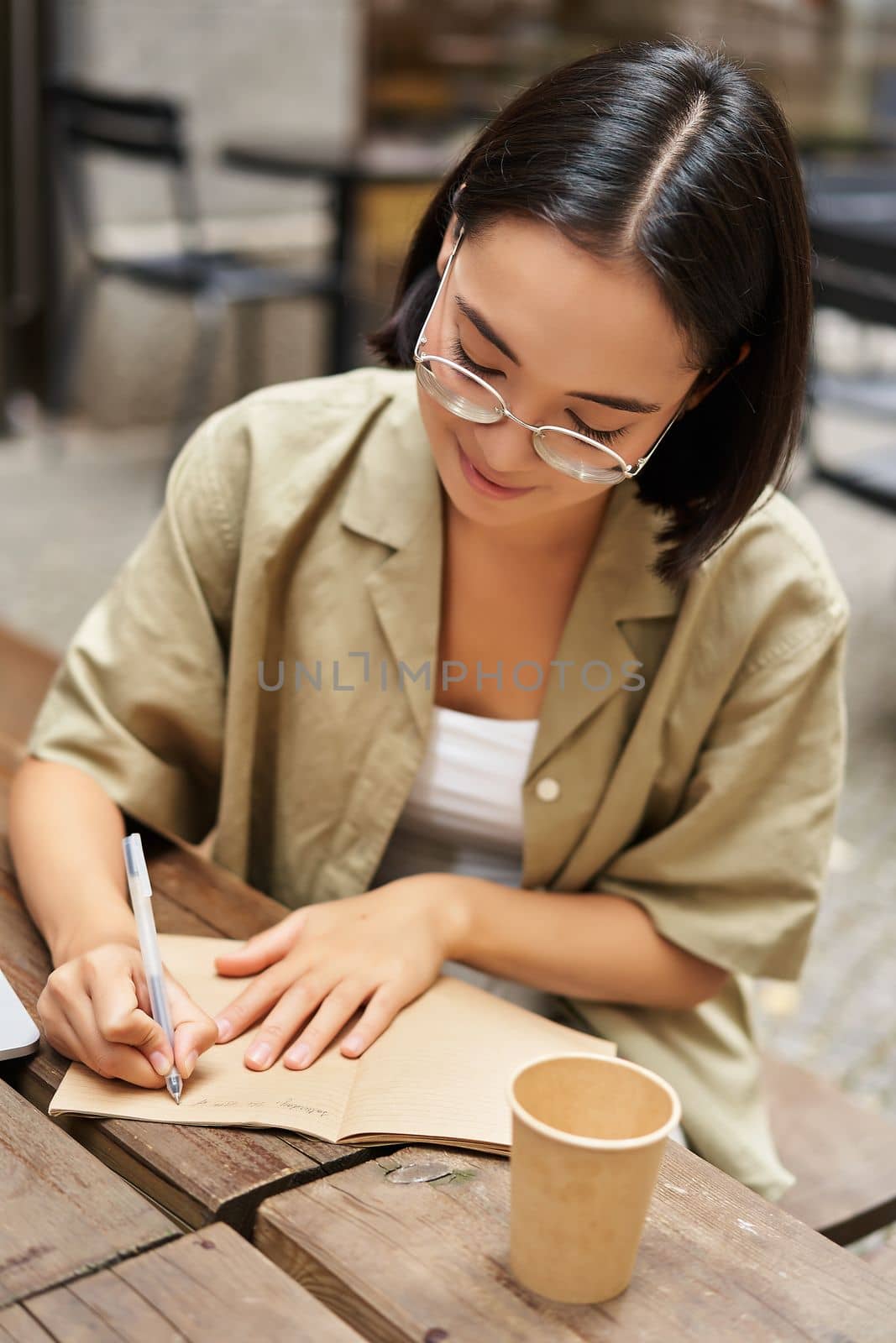 Vertical shot of young asian woman doing homework, making notes, writing something down, sitting in an outdoors cafe and drinking coffee.