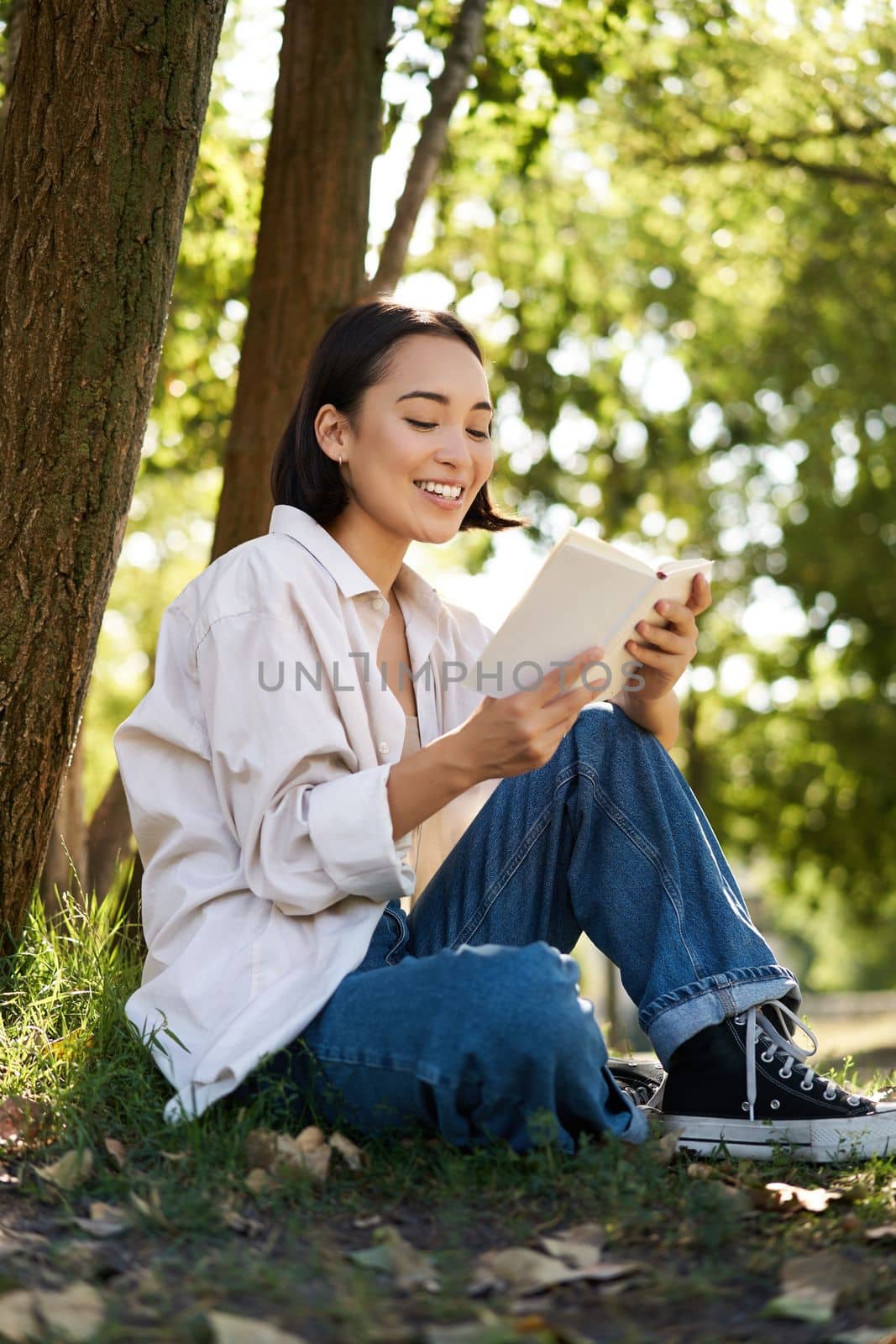 Beautiful young asian girl, student sits in park under tree and reading book, smiling, enjoying warm summer day outdoors.