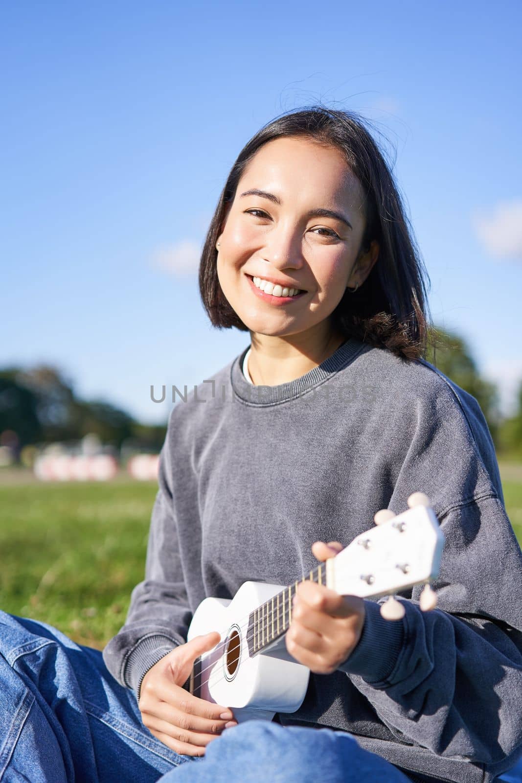 Portrait of cute smiling girl playing ukulele in park. Young woman with musical instrument sitting outdoors. Lifestyle and hobbies concept