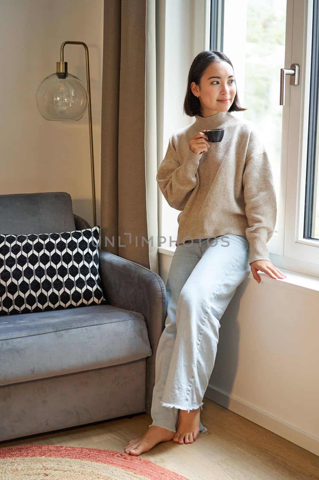 Vertical shot of beautiful woman sitting in her house, looking outside window and drinking coffee.