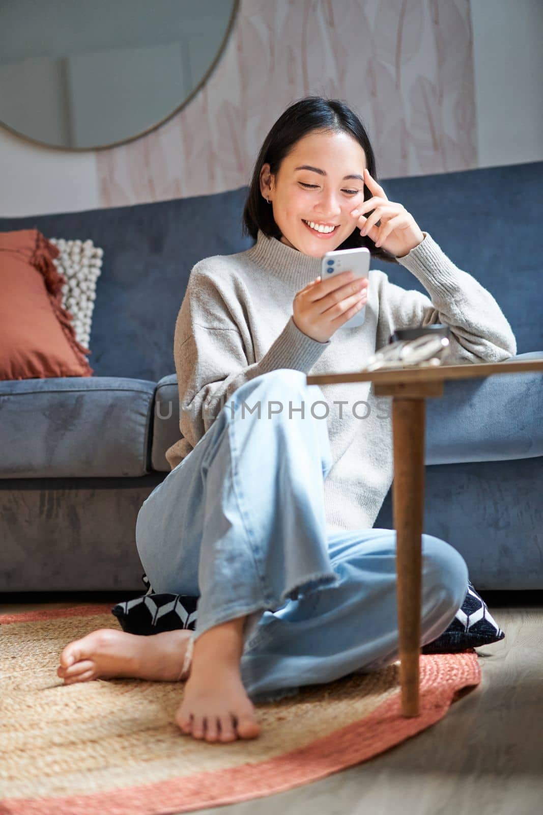 Vertical shot of stylish korean woman sitting on floor at home, using her mobile phone, holding smartphone and smiling.