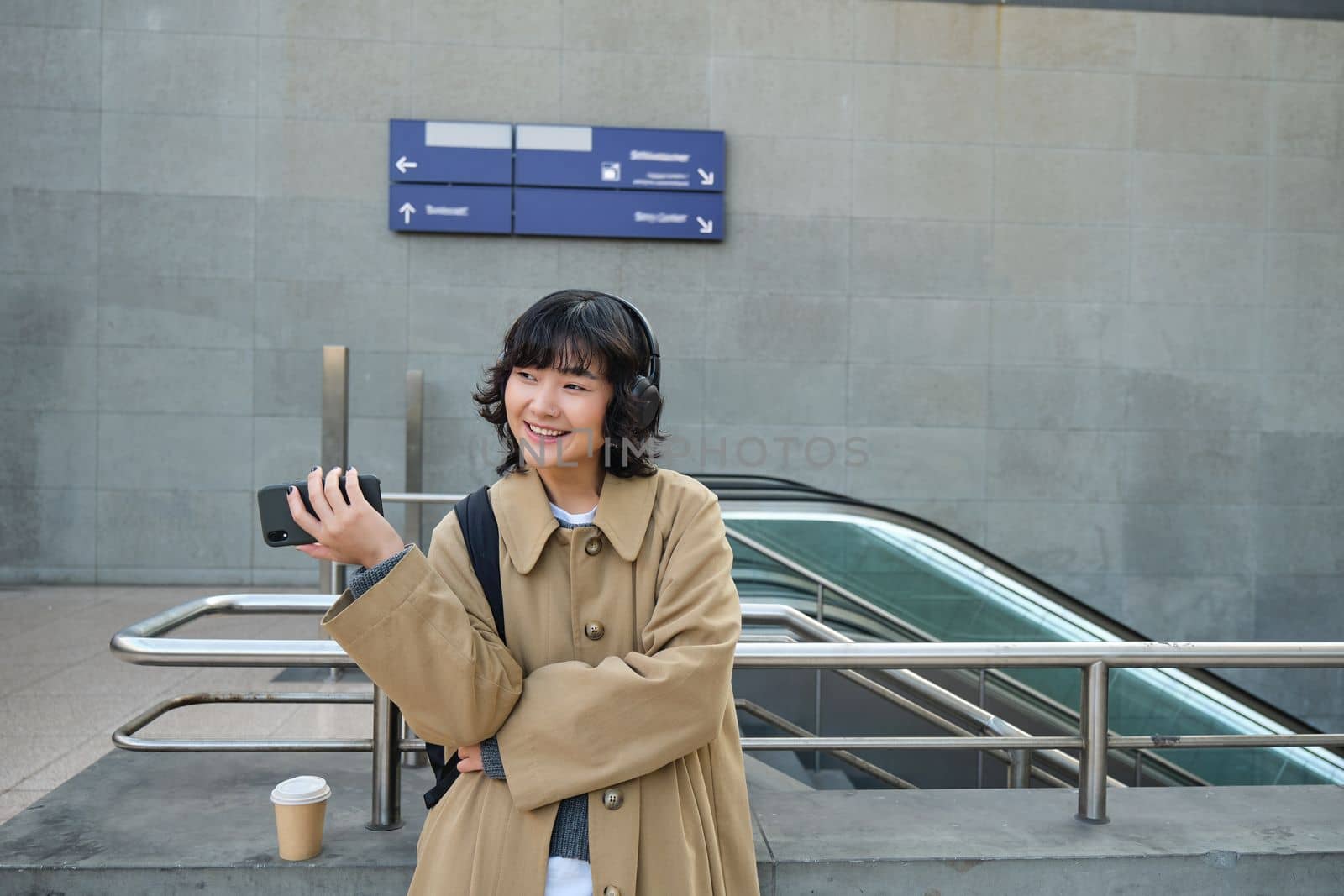 Beautiful brunette girl in trench coat, listens music in headphones and holds mobile phone, waits for someone on street and drinks coffee by Benzoix