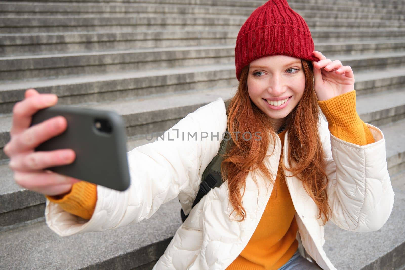 Stylish young girl in red hat, takes photos on smartphone camera, makes selfie as she sits on stairs near museum, posing for photo with app filter.