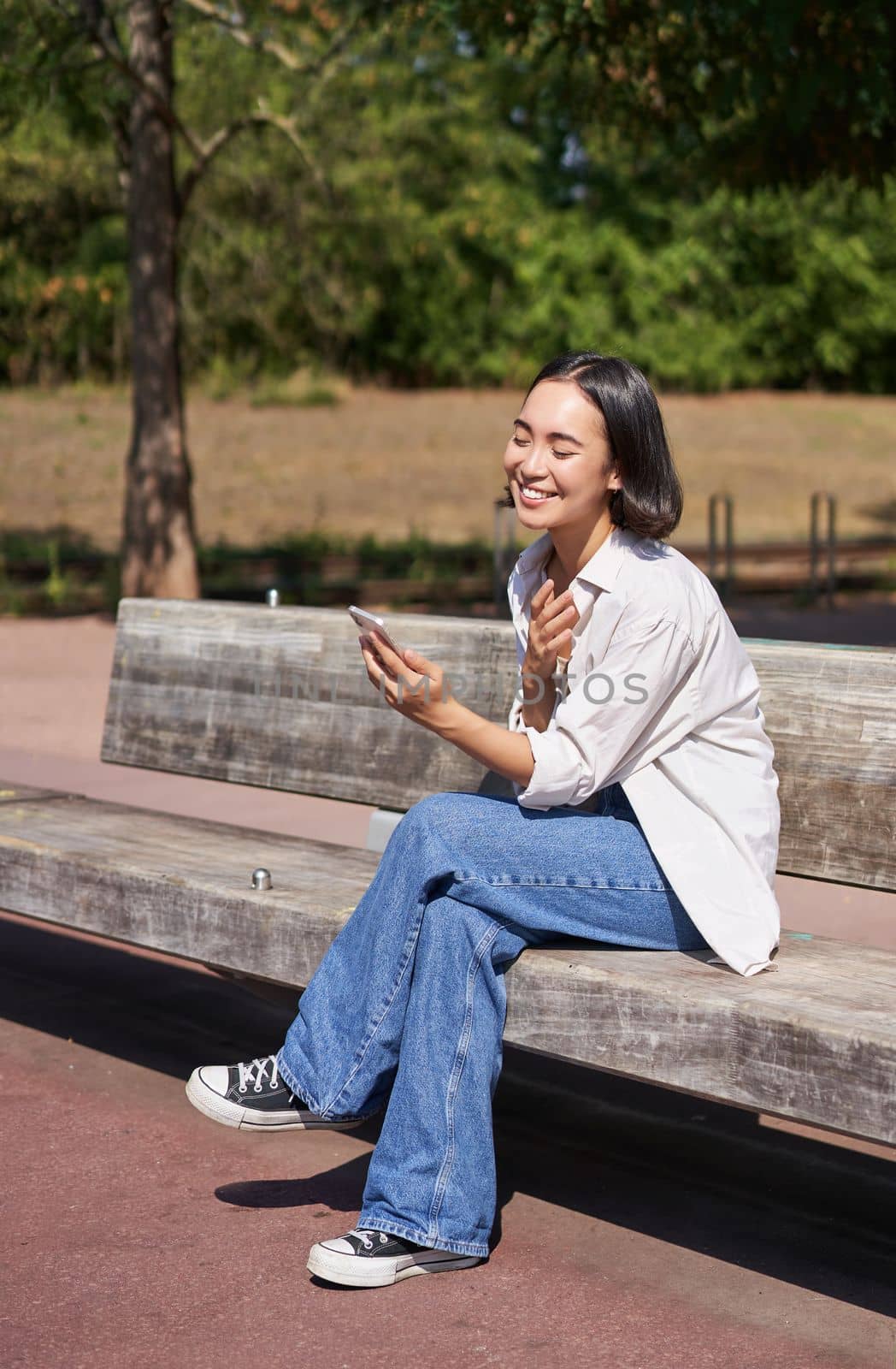 Stylish young asian girl, student sitting in park with smartphone, using telephone, waiting for someone while sitting on bench.