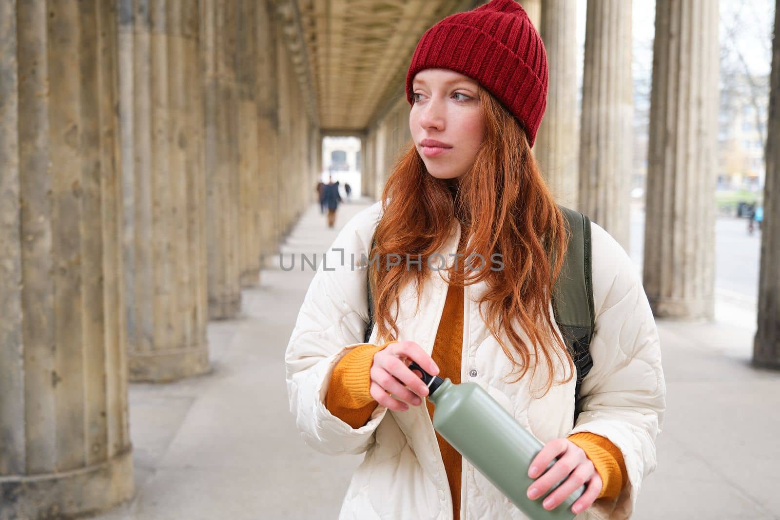 Young redhead girl holds thermos in hands, pours herself a hot drink while walking in city. Tourist takes break, opens flask for refreshment beverage.