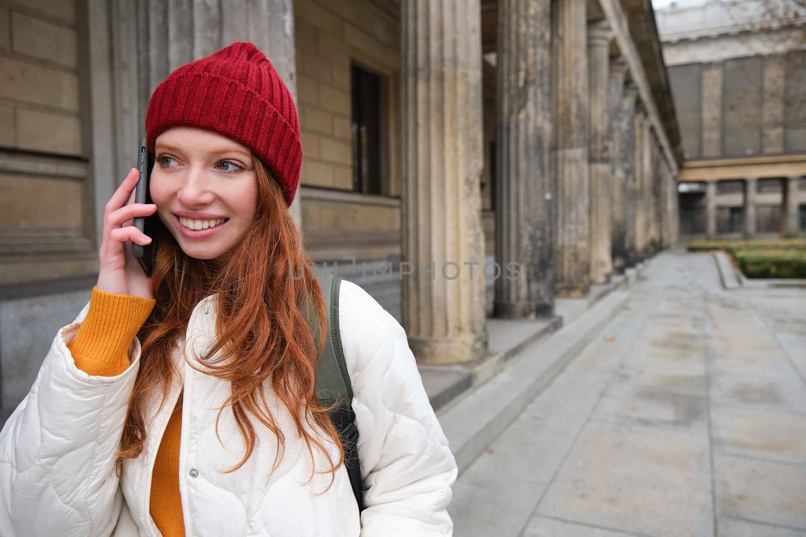 Smiling redhead female tourist talks on mobile phone and walks around city. Happy student in red hat calls friend, stands on street and uses smartphone by Benzoix