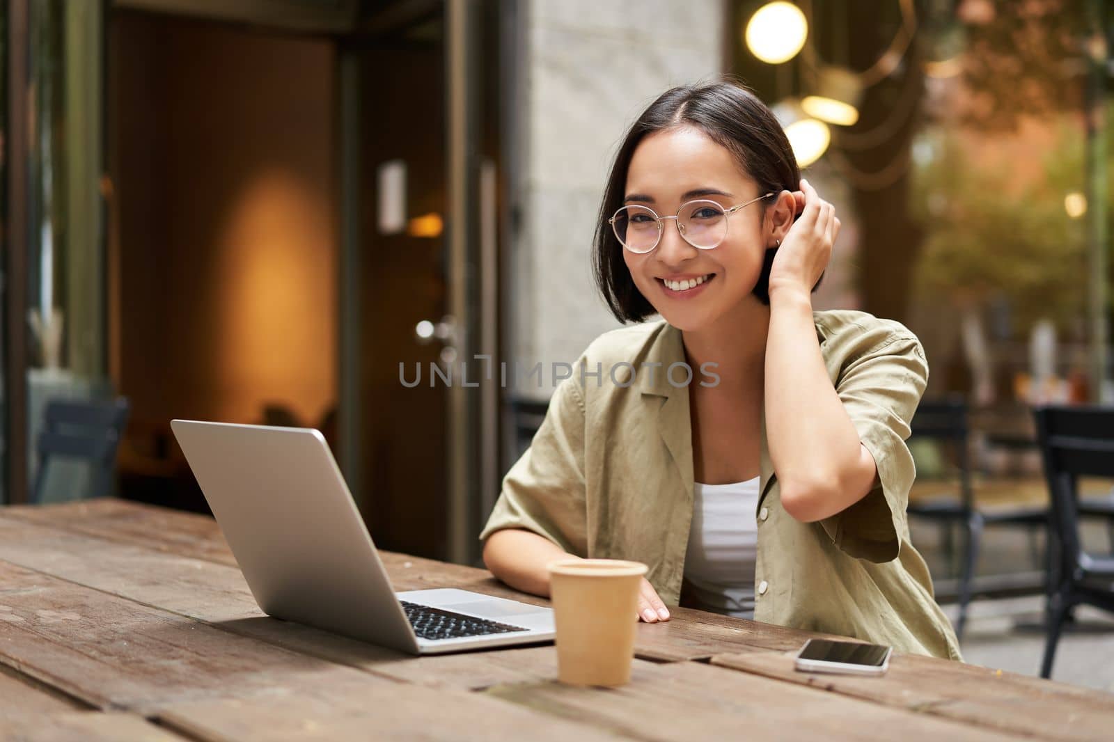 Portrait of smiling girl in glasses, sitting with laptop in outdoor cafe, drinking coffee and working remotely, studying online.