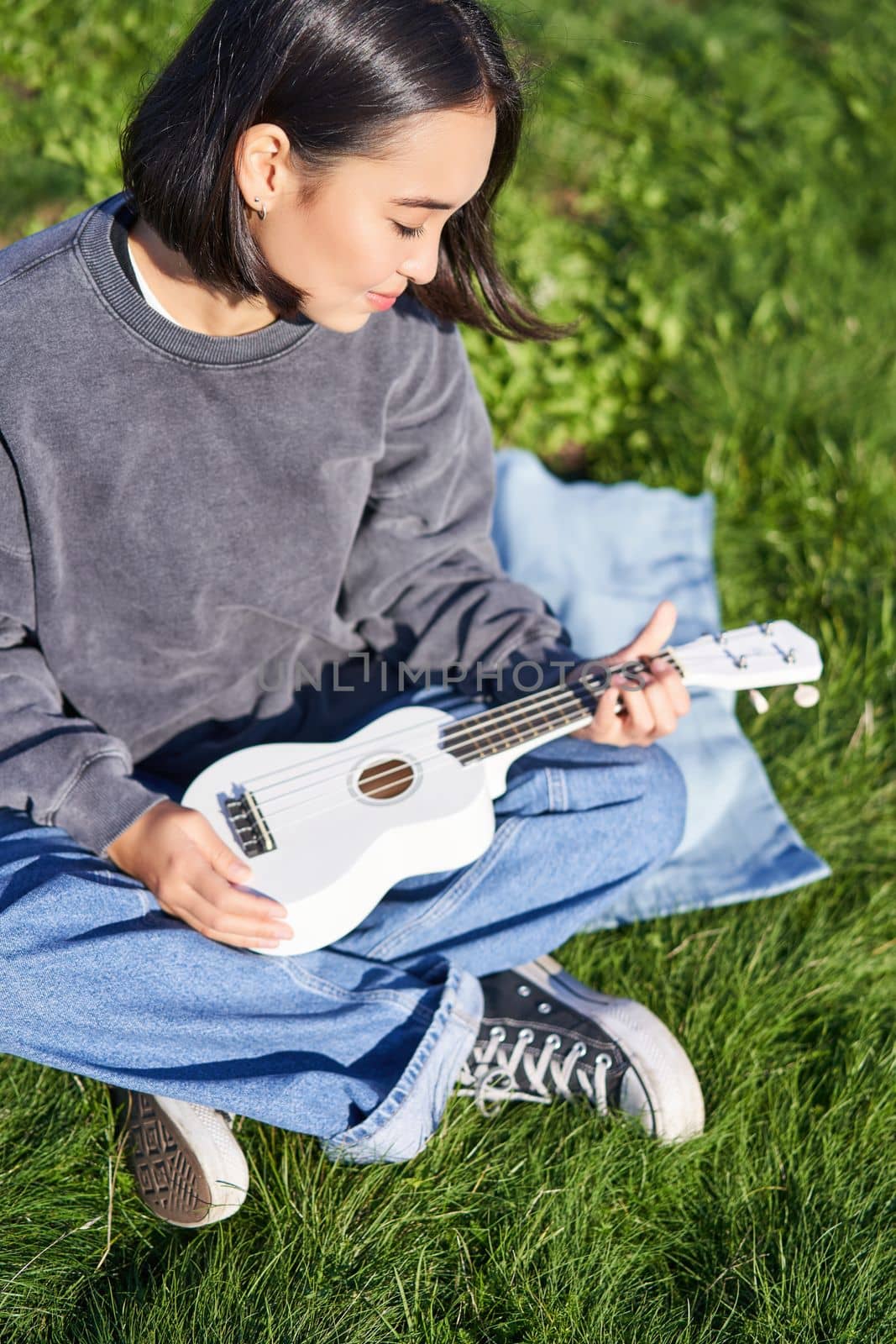 Vertical shot of girl musician, looking with care at her white ukulele guitar, playing music in park, sitting on grass.
