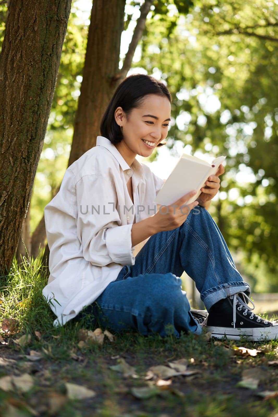 Vertical shot of happy asian woman relaxing outdoors in park, reading her book and sitting under tree shade on warm sunny day.