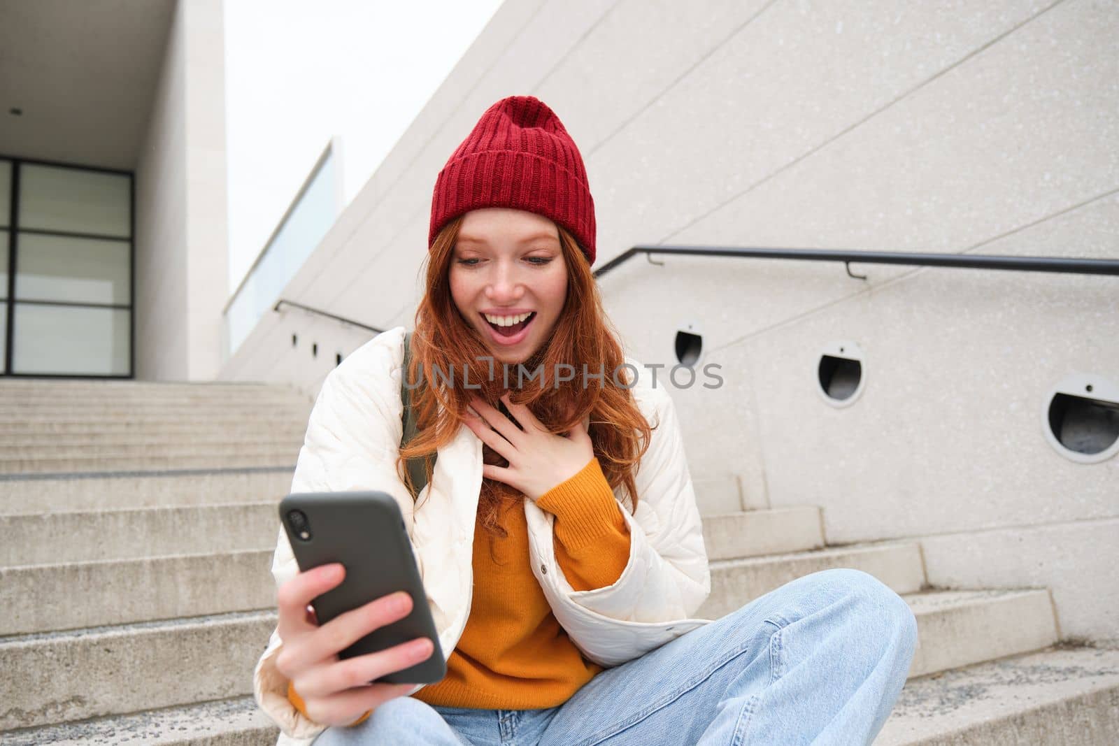 Image of redhead girl with surprised face expression, looks amazed at her smartphone, reads great news on phone, sits on street stairs.