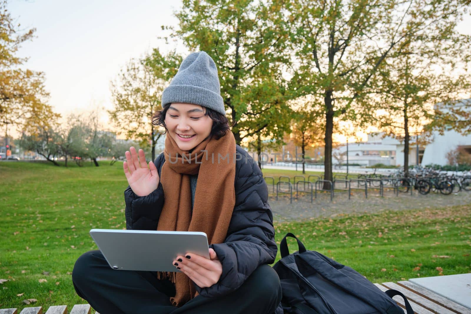 Portrait of young asian girl sits in warm clothes in park, waves hand at tablet, video chats outdoors, says hello while on call by Benzoix