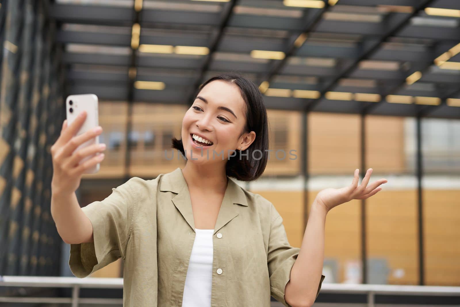 Happy asian girl shows something behind her during video call, demonstrating smth, standing on street by Benzoix