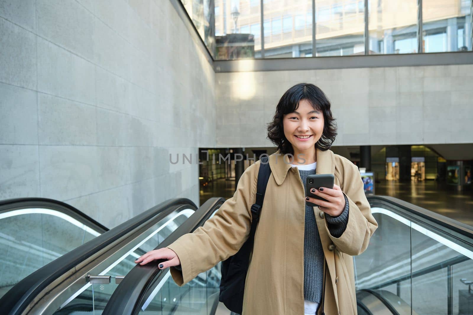 Technology and people concept. Smiling brunette woman travels around city, holds smartphone and laughs, uses mobile phone on her way home, stands on escalator.