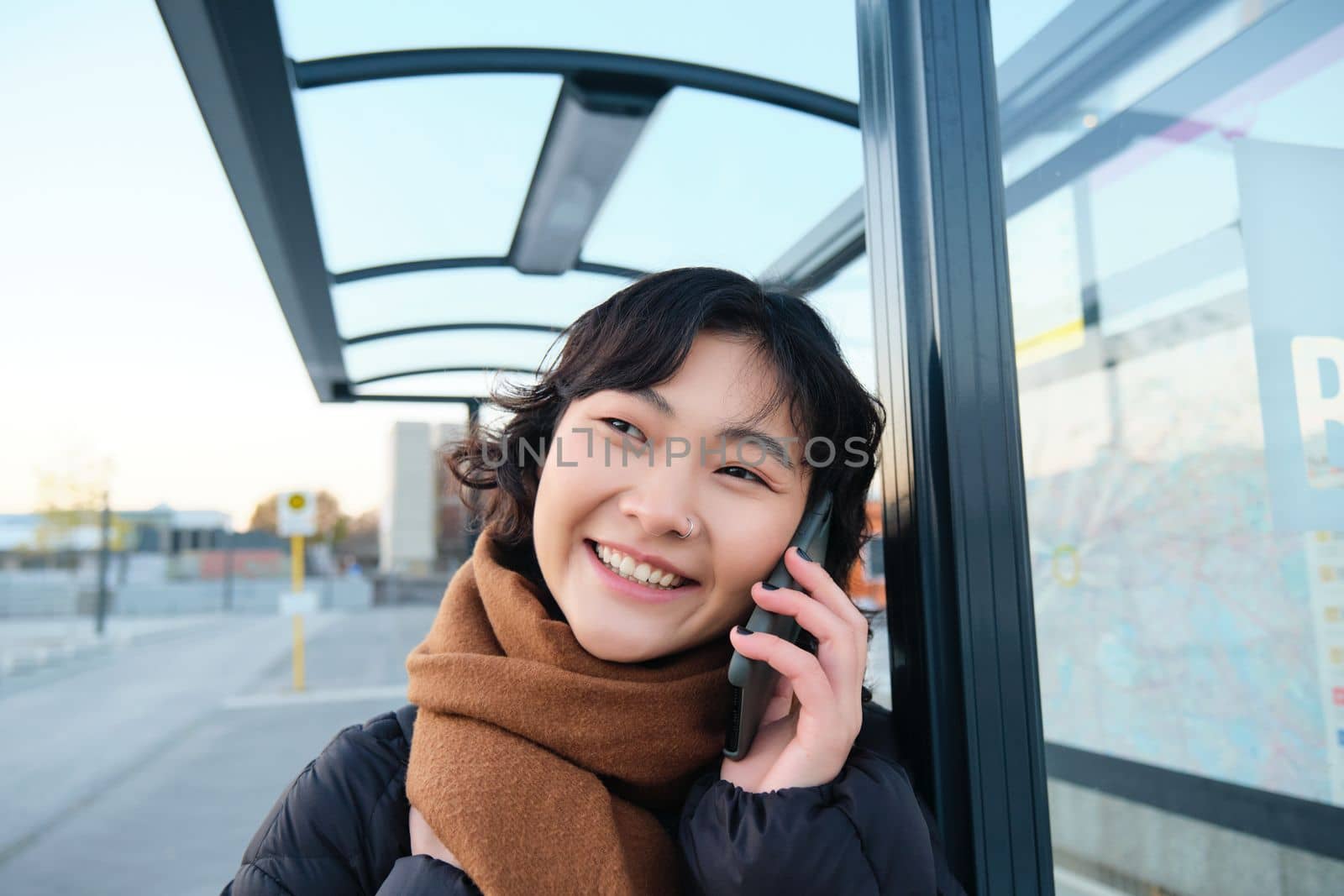 Close up of cute Korean woman, making a phone call, talking and smiling on telephone, standing in winter jacket on bus stop, waiting for her transport to arrive.