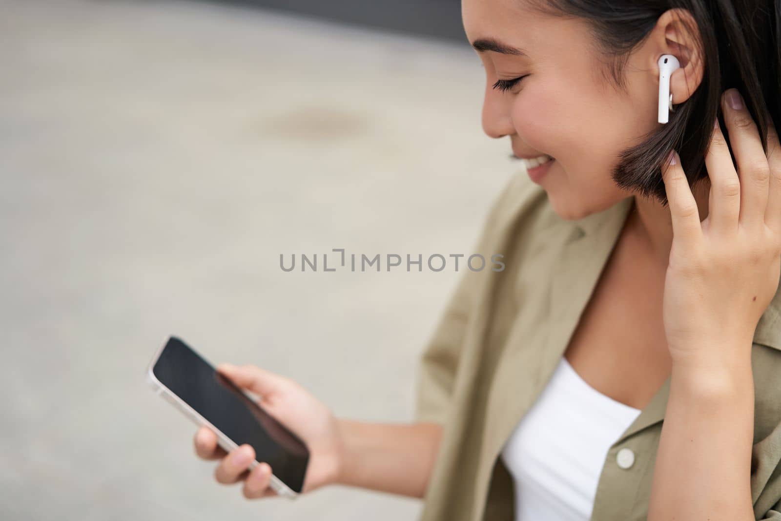 Close up portrait of asian girl, looking at mobile screen, listening music in headphones. Woman with earphones walks on street.