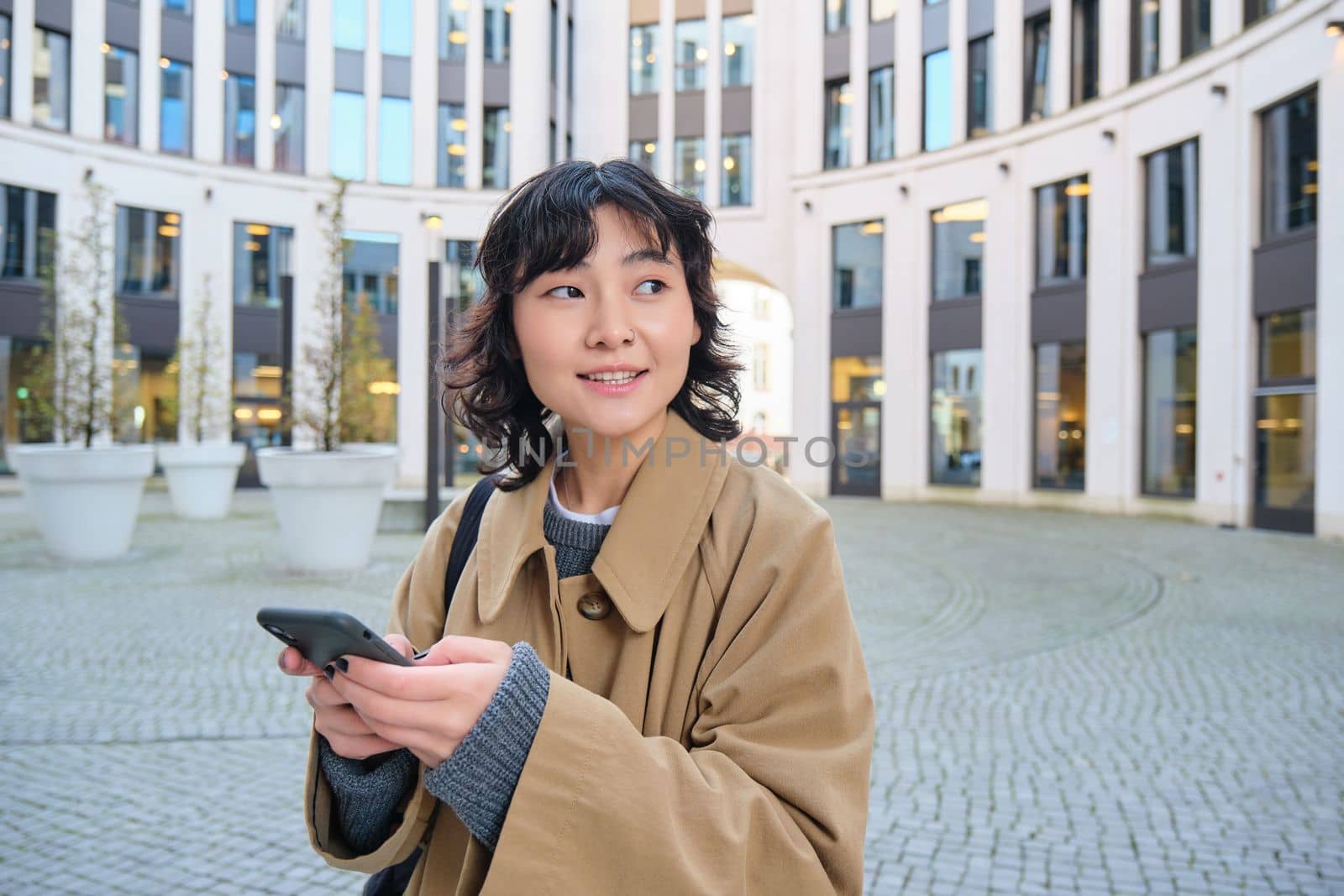 Stylish korean girl in headphones, listens music and uses mobile phone, stands in city centre, waits for someone on street and writes text message on her smartphone.