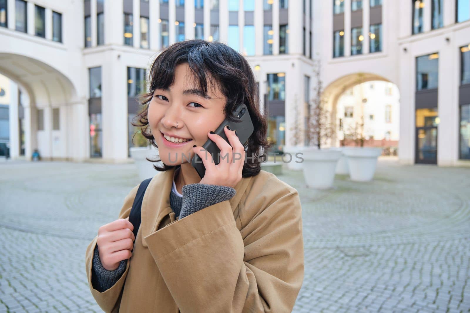 Cellular technology. Young korean woman talks on mobile phone, makes a phone call on her way home, walks down street, city centre, has telephone conversation.