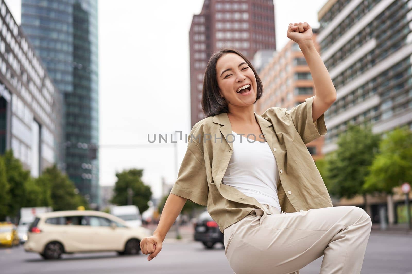 Portrait of happy asian woman, dancing and feeling joy, triumphing, raising hand up in victory gesture, celebrating on streets by Benzoix