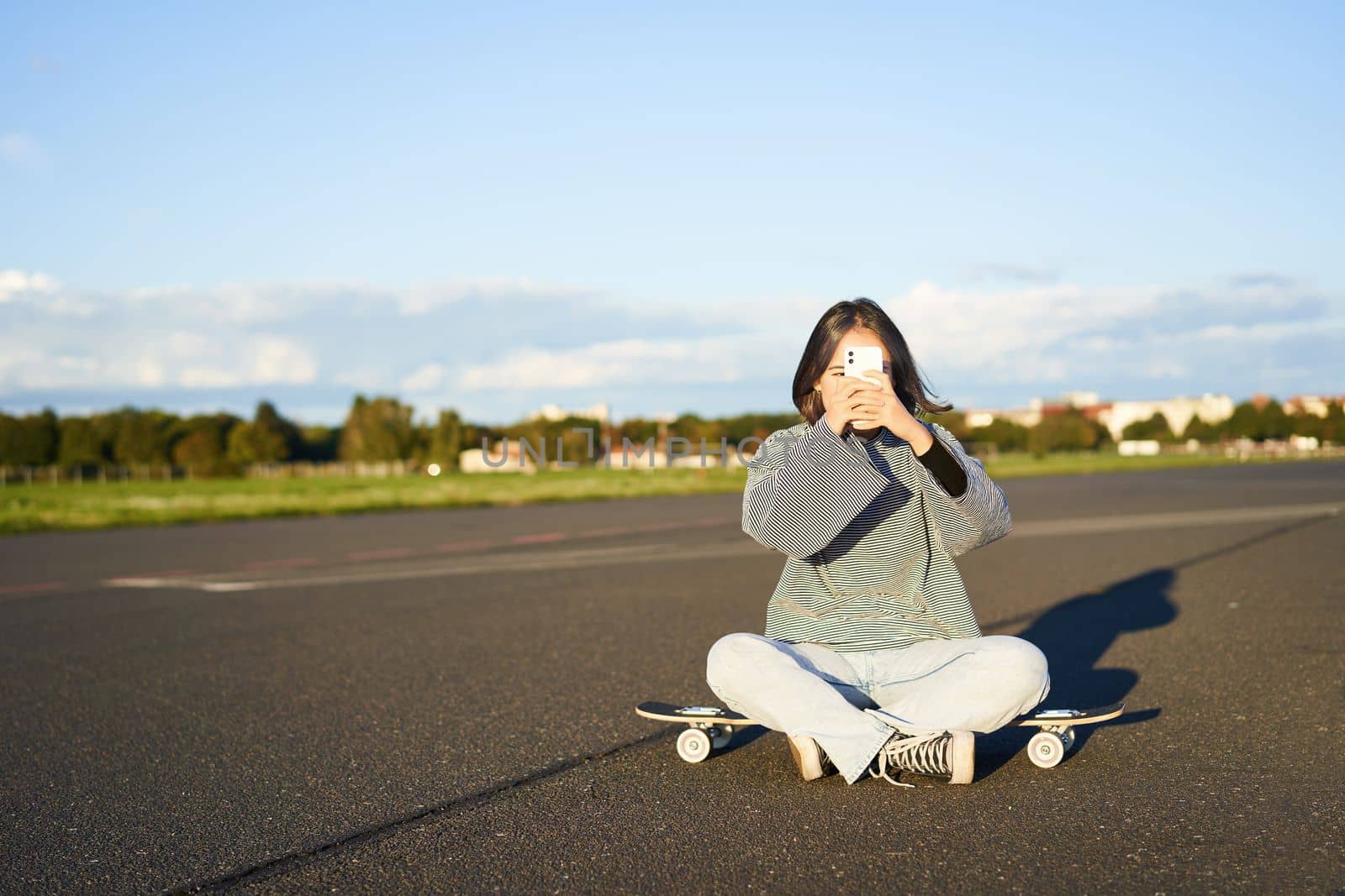 Hipster teen girl sitting on her skateboard, taking photos on smartphone. Asian woman skater sits on longboard and photographing on mobile phone by Benzoix