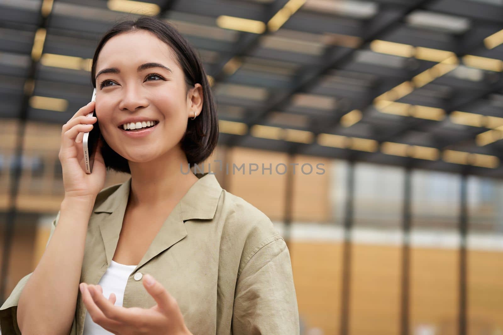 Young asian woman smiles, talks on mobile phone and walks in city centre, poses outdoors.