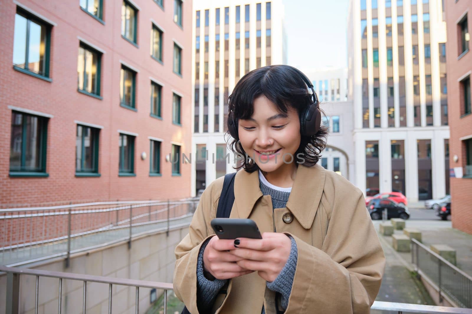 Portrait of cute girl tourist, korean woman in headphones, looks at mobile phone, uses smartphone app, map or text messages, listens music in headphones by Benzoix