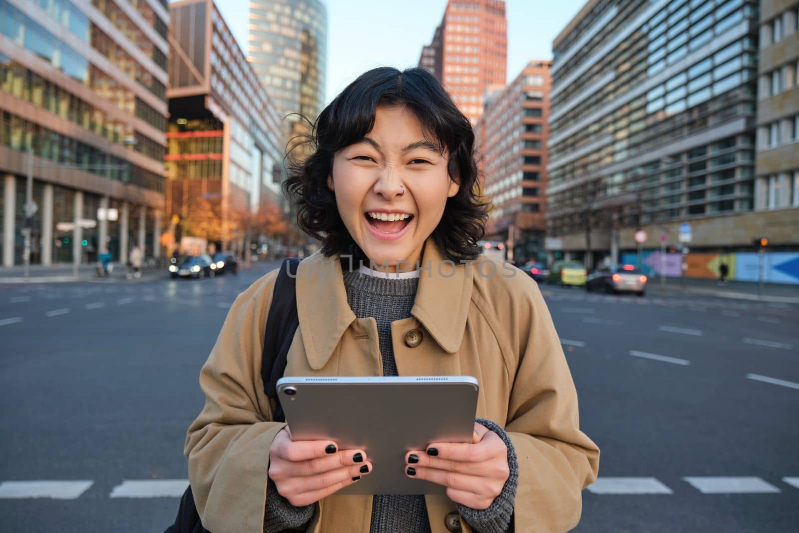 Enthusiastic asian girl with digital tablet, smiling and looking amazed, happy with something good, holding digital tablet, standing on street.