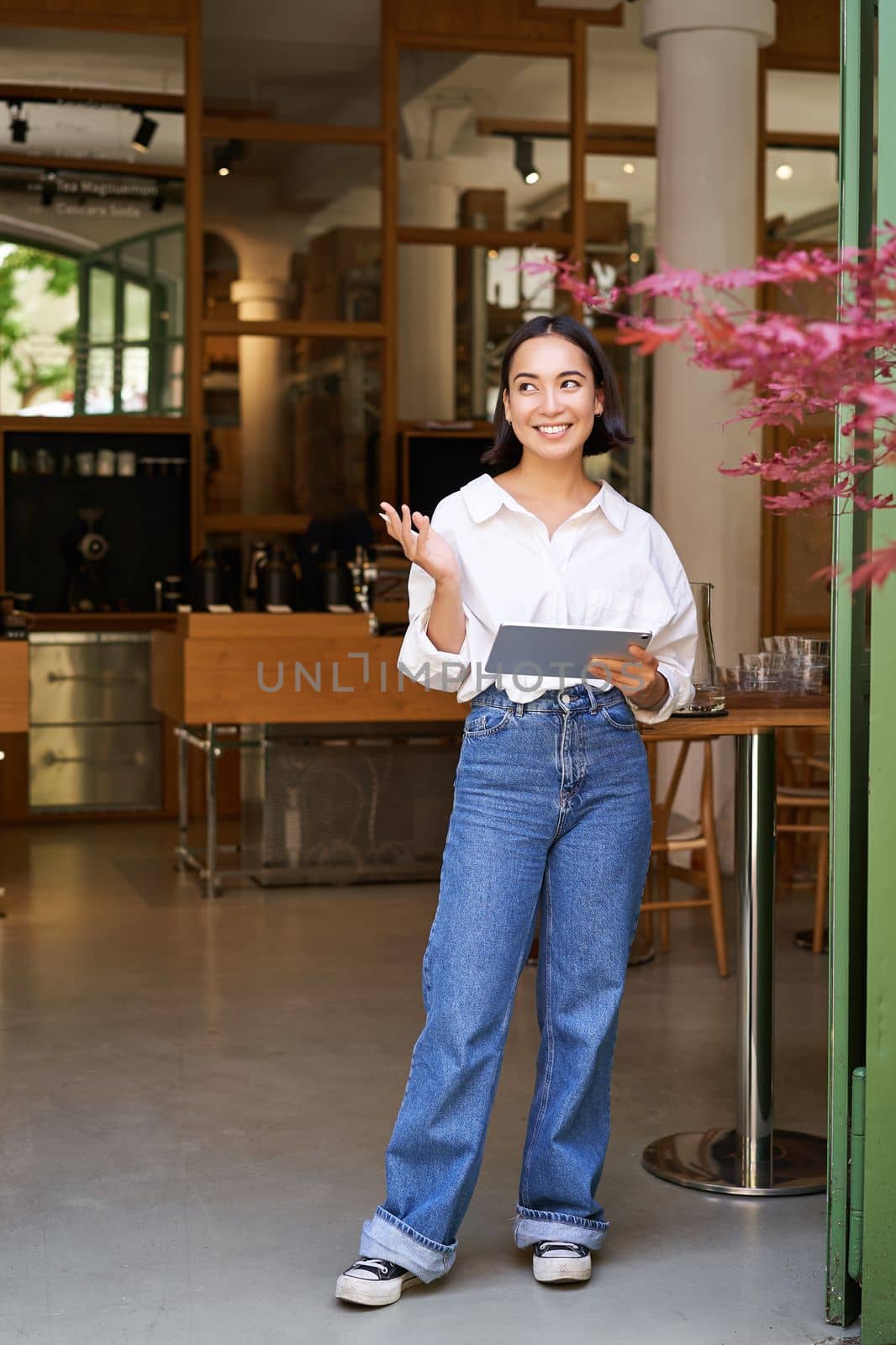 Vertical shot of smiling asian businesswoman, waitress standing in front of cafe entrance, inviting guests in restaurant with tablet.