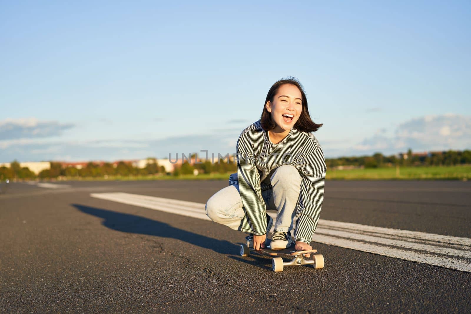 Freedom and happiness. Happy asian girl riding her longboard on an empty sunny road, laughing and smiling, skateboarding by Benzoix