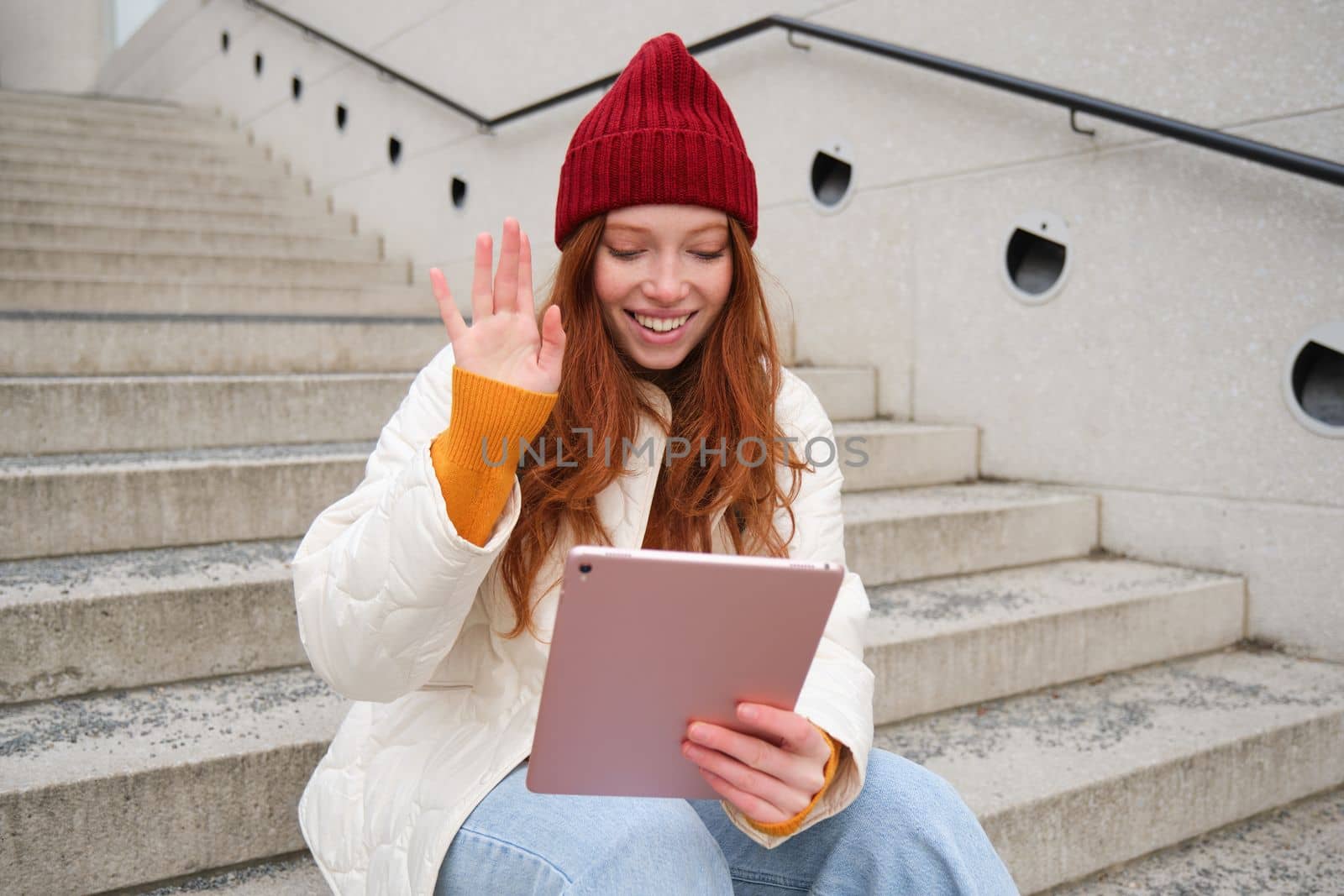 Joyful redhead teen girl, student with digital tablet, says hello, waves hand at gadget camera, connects to video chat, talks to friend in application by Benzoix