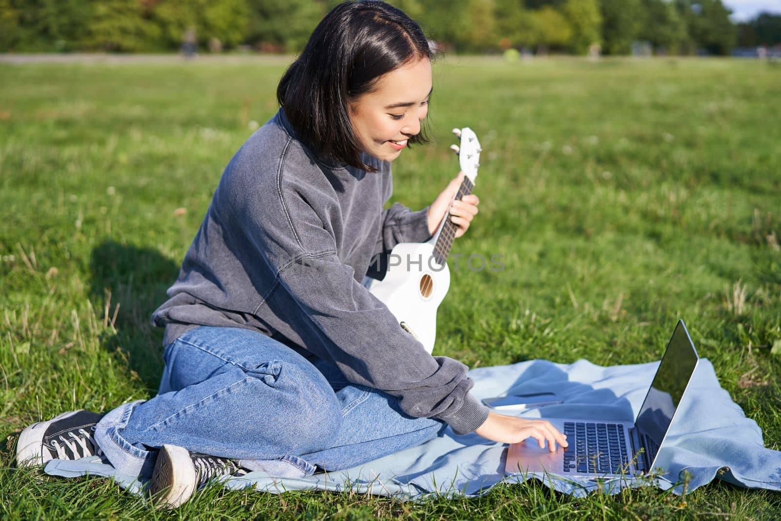 Smiling asian girl learns how to play ukulele via laptop, online video tutorials, sitting on grass in park with musical instrument.