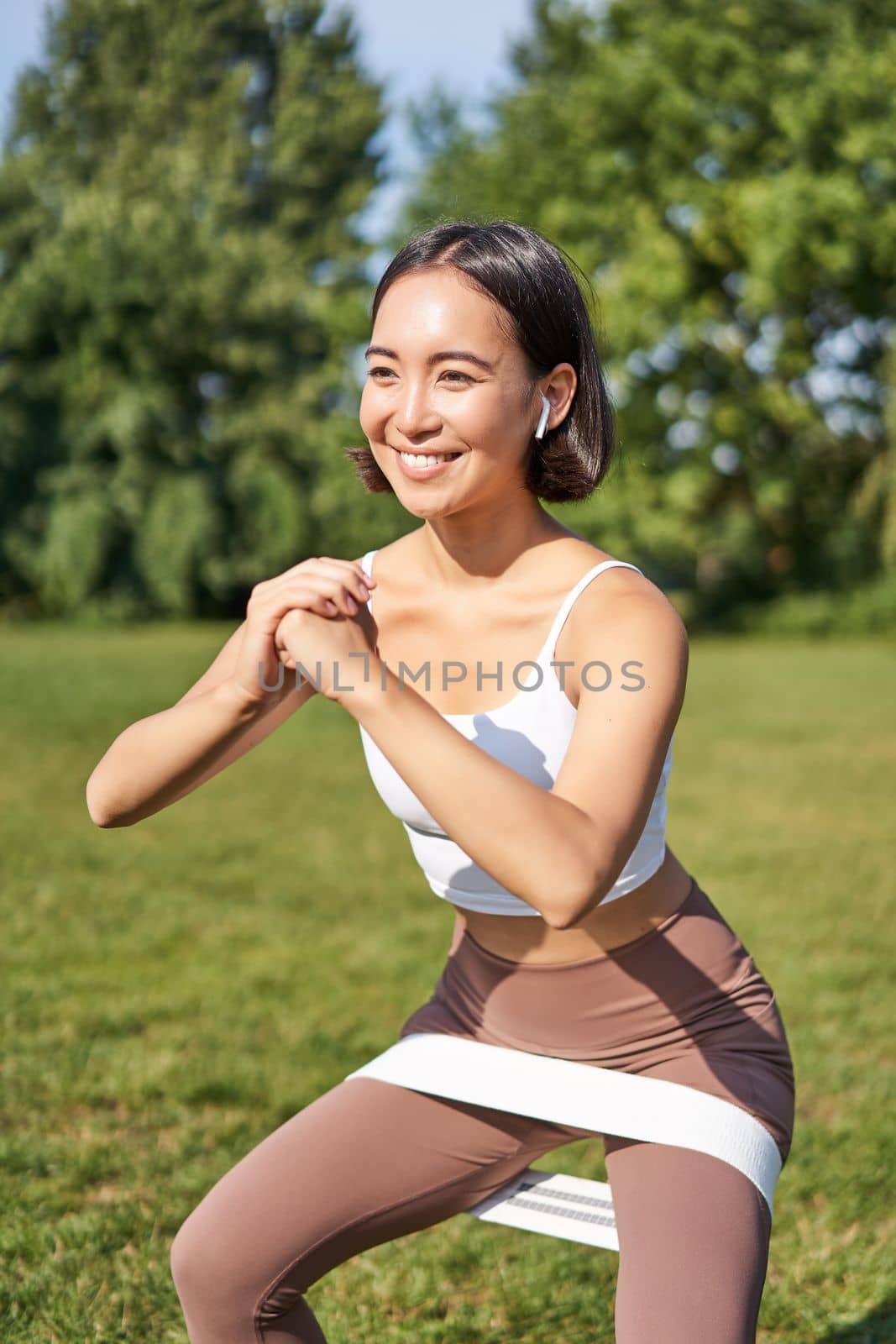 Vertical shot of young fit woman does squats in park, using stretching band on legs, smiling pleased while workout.