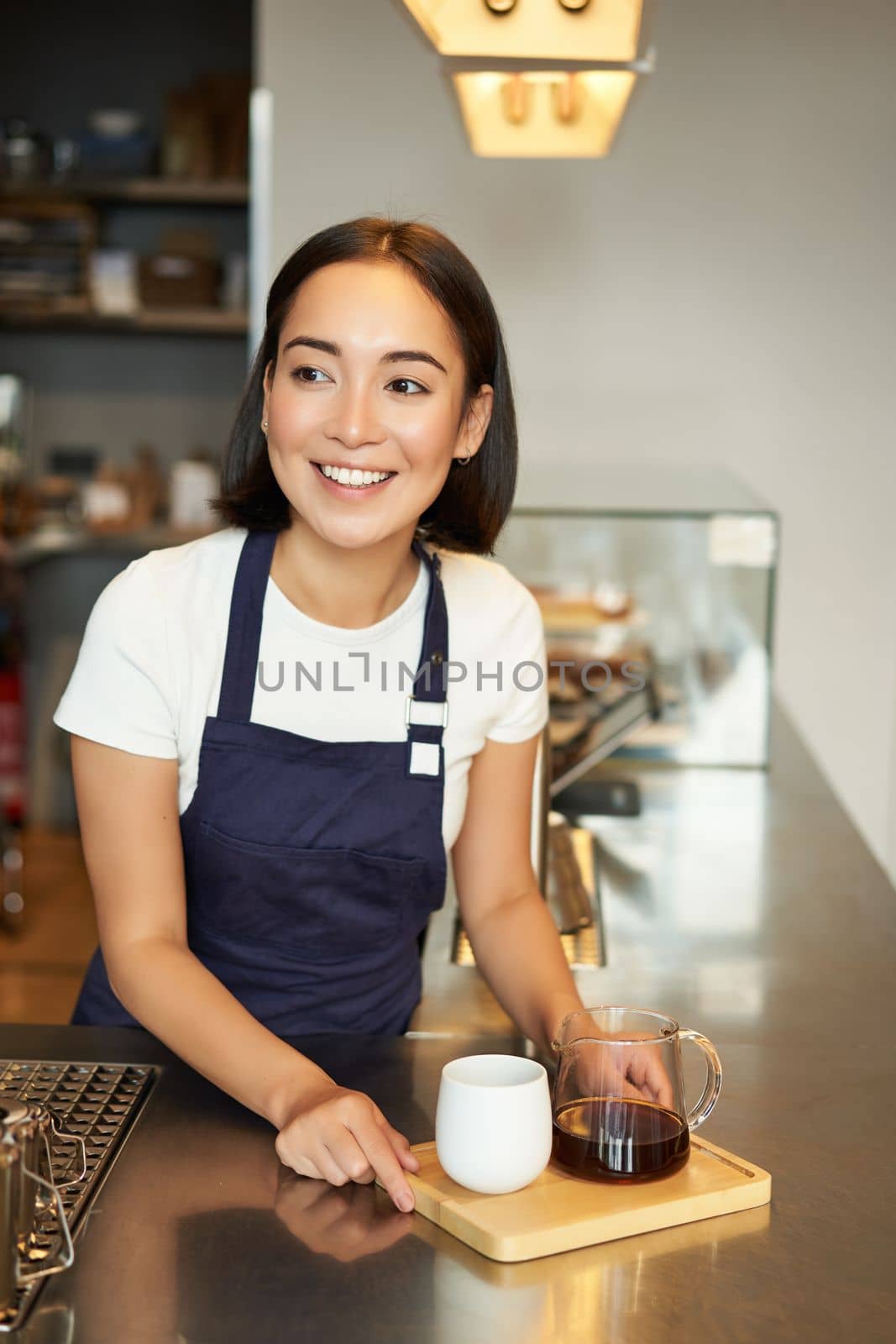 Vertical shot of smiling girl barista serving coffee, making batch brew, filter for client in cafe, wearing blue apron behind counter.