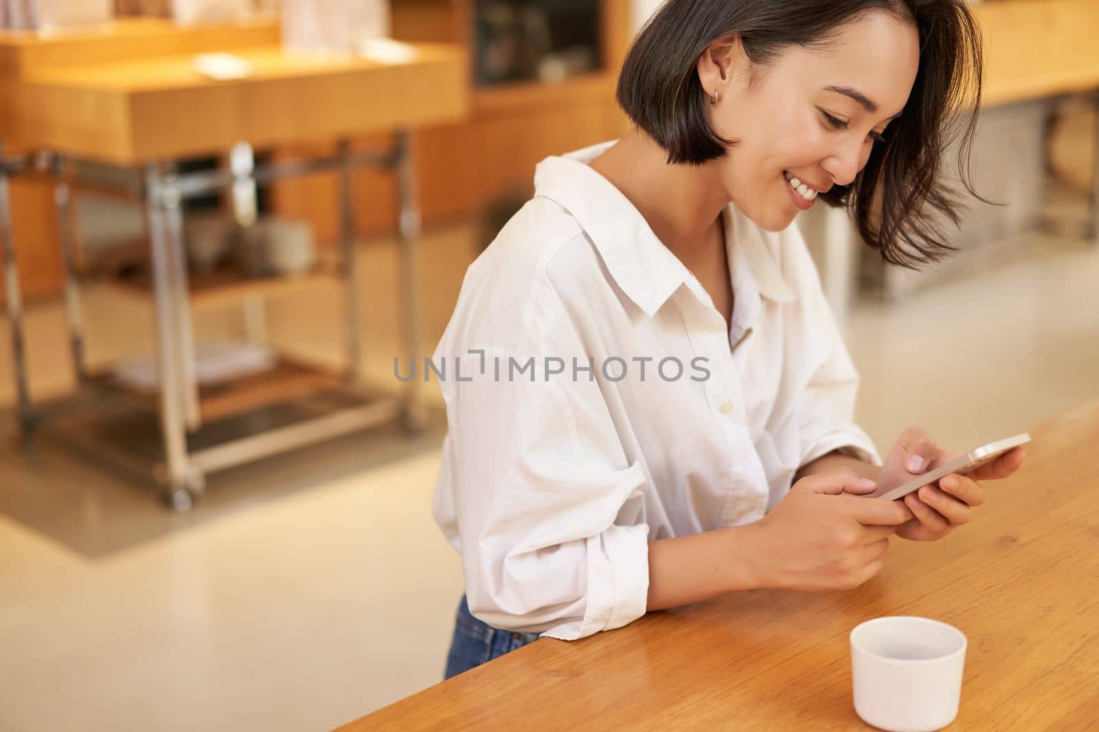 Portrait of young asian woman, sitting in cafe, holding smartphone, chatting and messaging while drinking coffee.