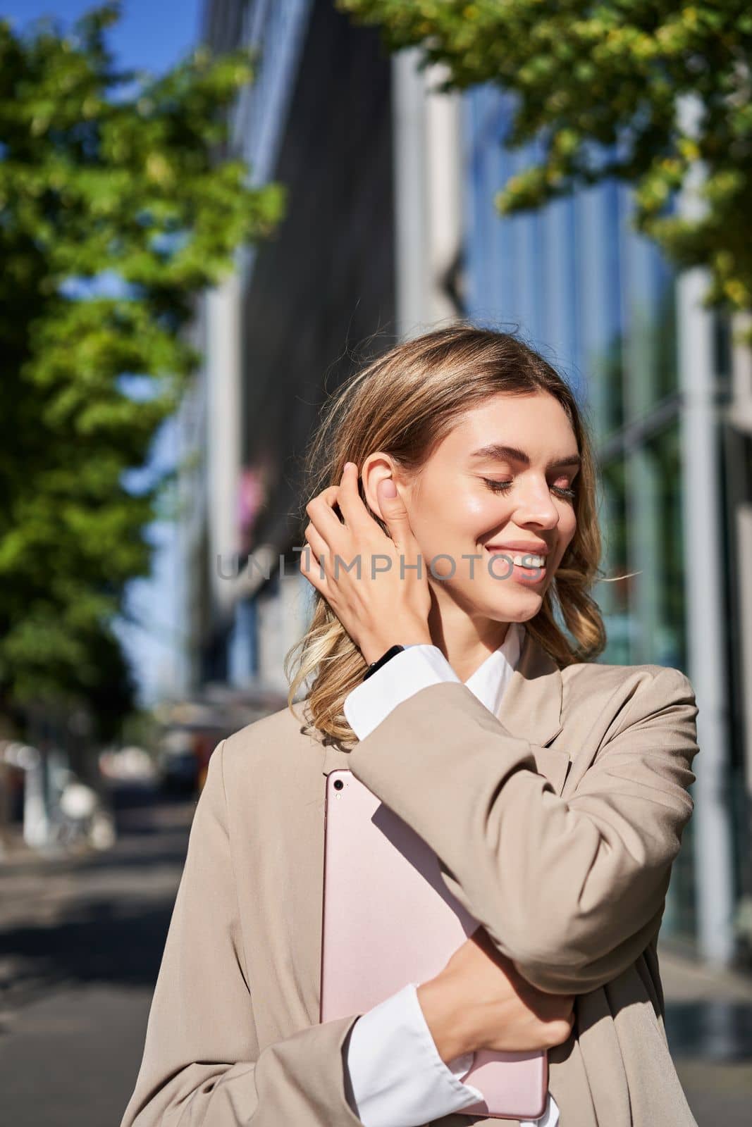 Vertical portrait of corporate woman holding digital tablet, posing on street, adjusting her hair and smiling.