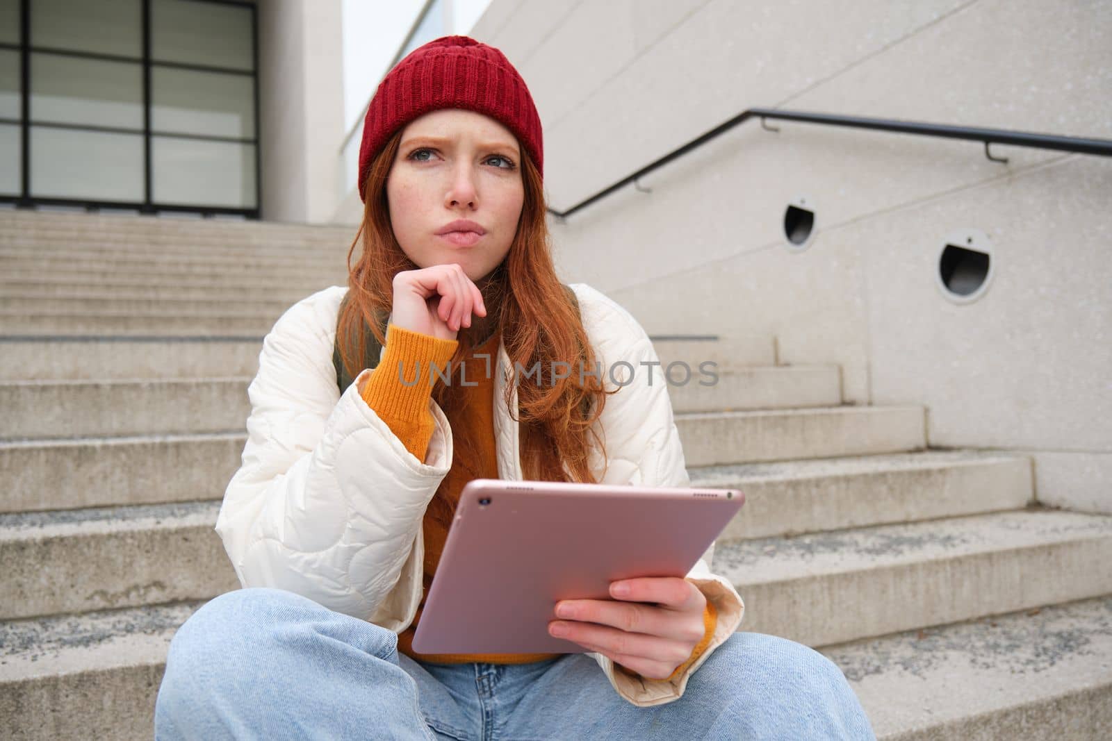 Portrait of troubled redhead girl, college student with thoughtful face, sits on street stairs, holds digital tablet, thinks how to reply on message.