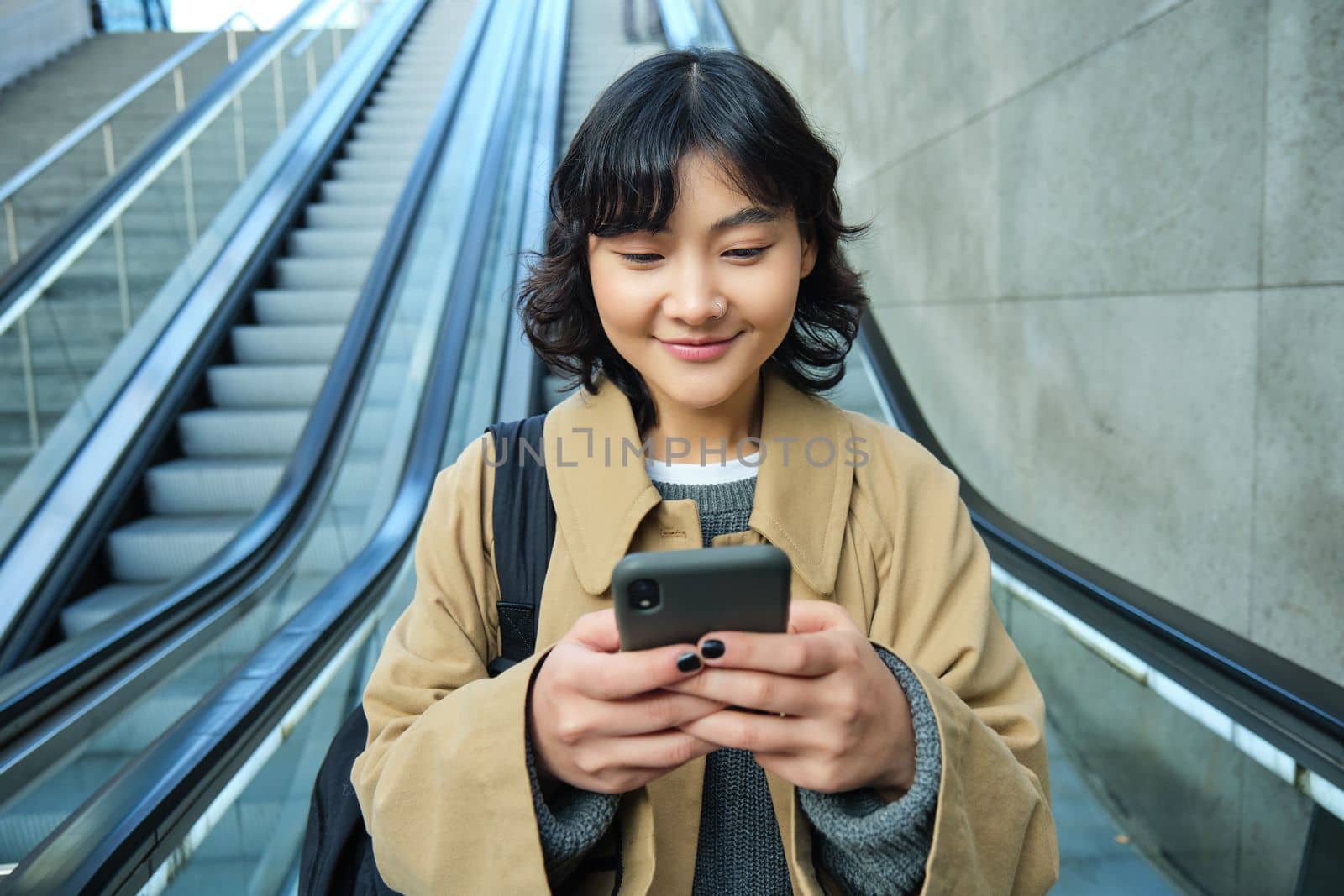 Portrait of beautiful asian girl, student goes down escalator in city, looks at mobile phone, uses telephone, map application, commutes somewhere in town.