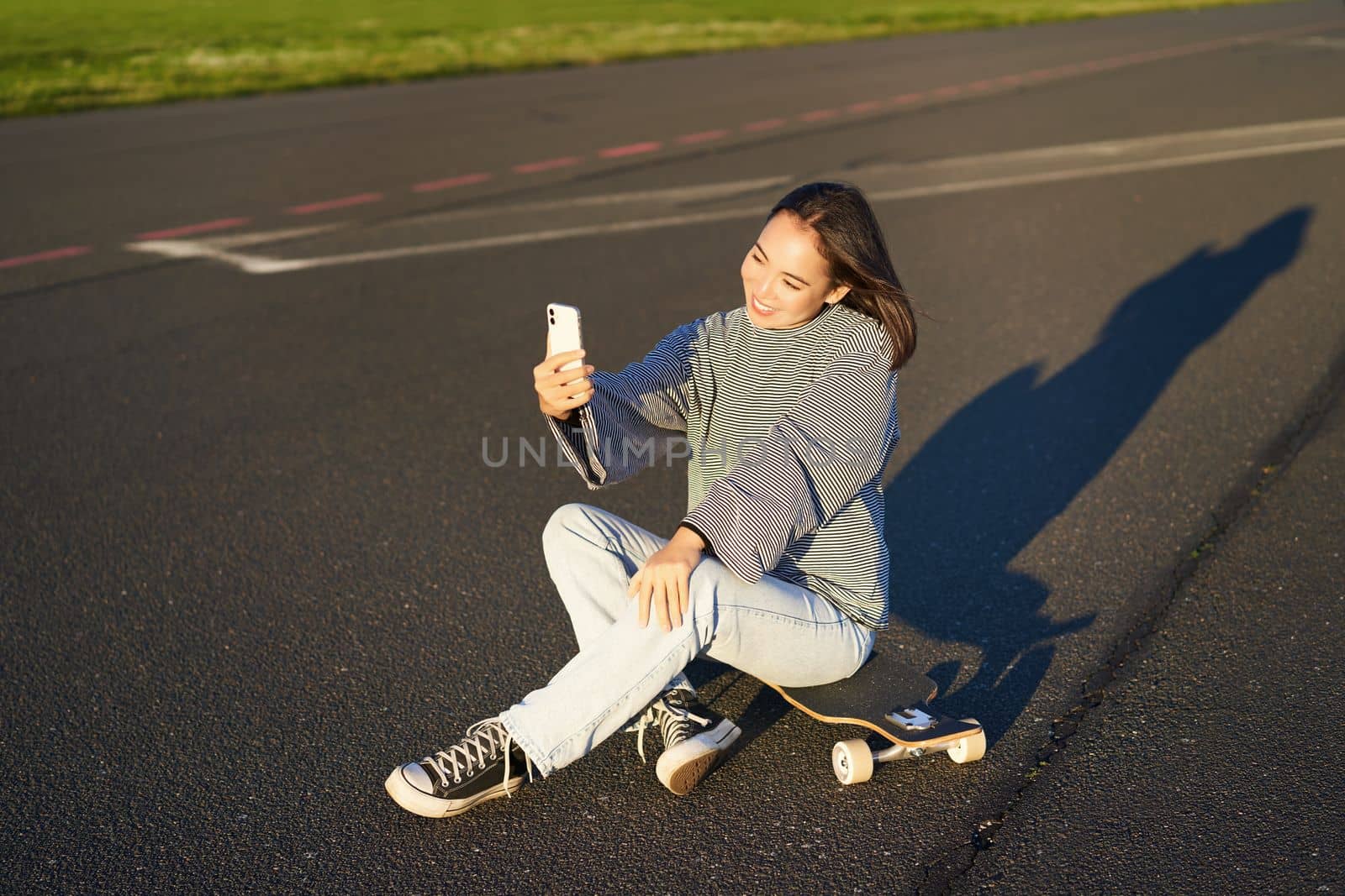 Beautiful korean girl takes selfie on smartphone, takes photo with her skateboard, enoys sunny day outdoors.