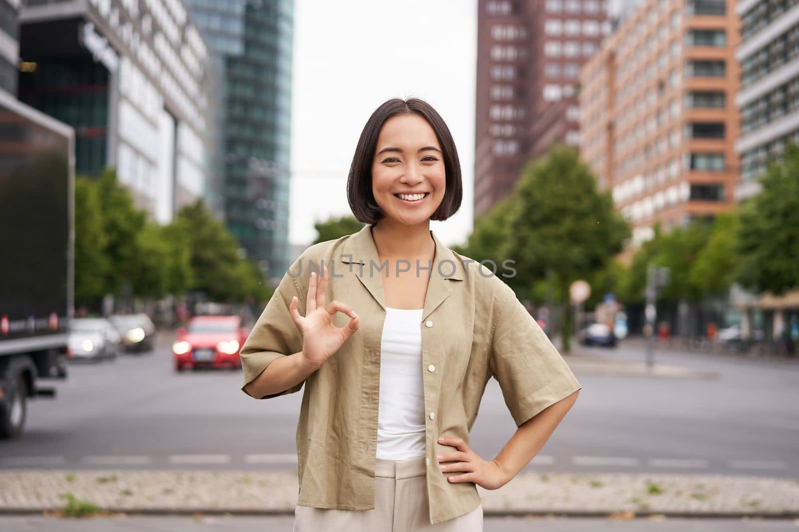 Enthusiastic city girl, shows okay gesture in approval, looking upbeat, say yes, approves and agrees, stands on street by Benzoix