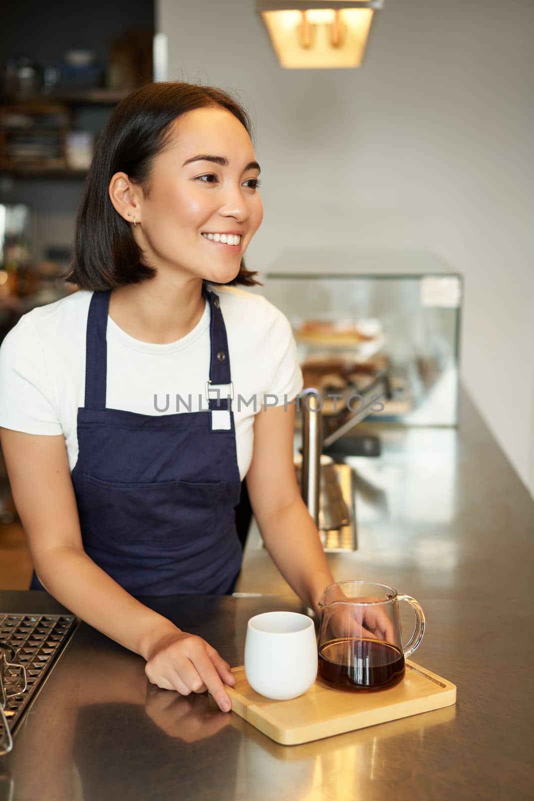 Vertical shot of smiling girl barista serving coffee, making batch brew, filter for client in cafe, wearing blue apron behind counter.