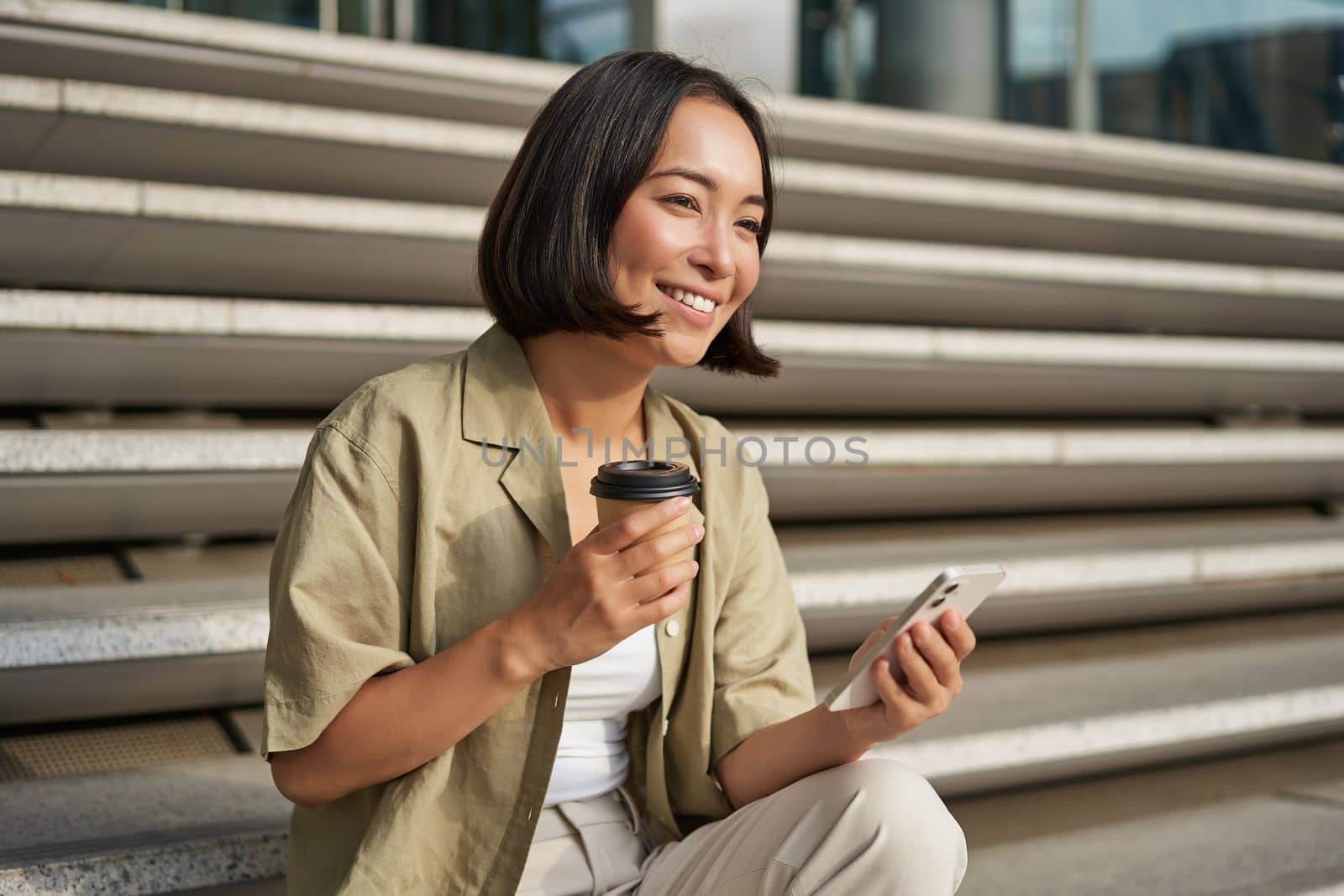 Portrait of asian woman with smartphone, drinks coffee and watches videos on mobile phone. Girl with telephone sits on stairs outdoors.