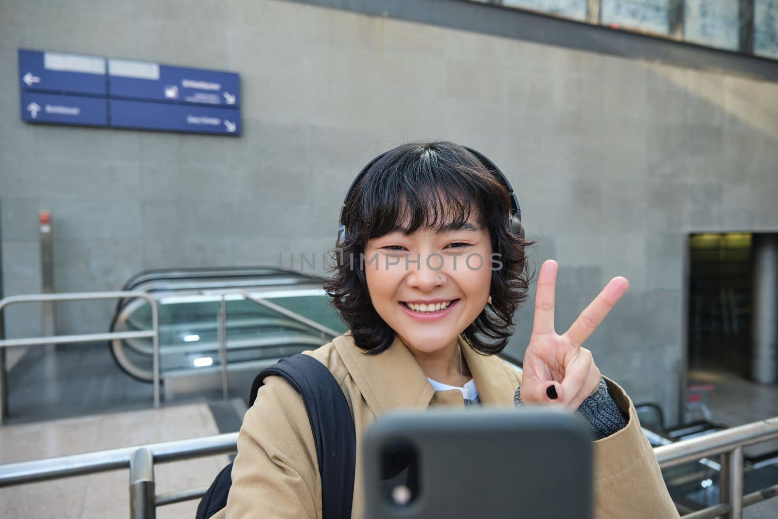 Cute and stylish Korean girl, wears headphones, takes selfie on smartphone, tourist records video or makes a photo, stands on street.