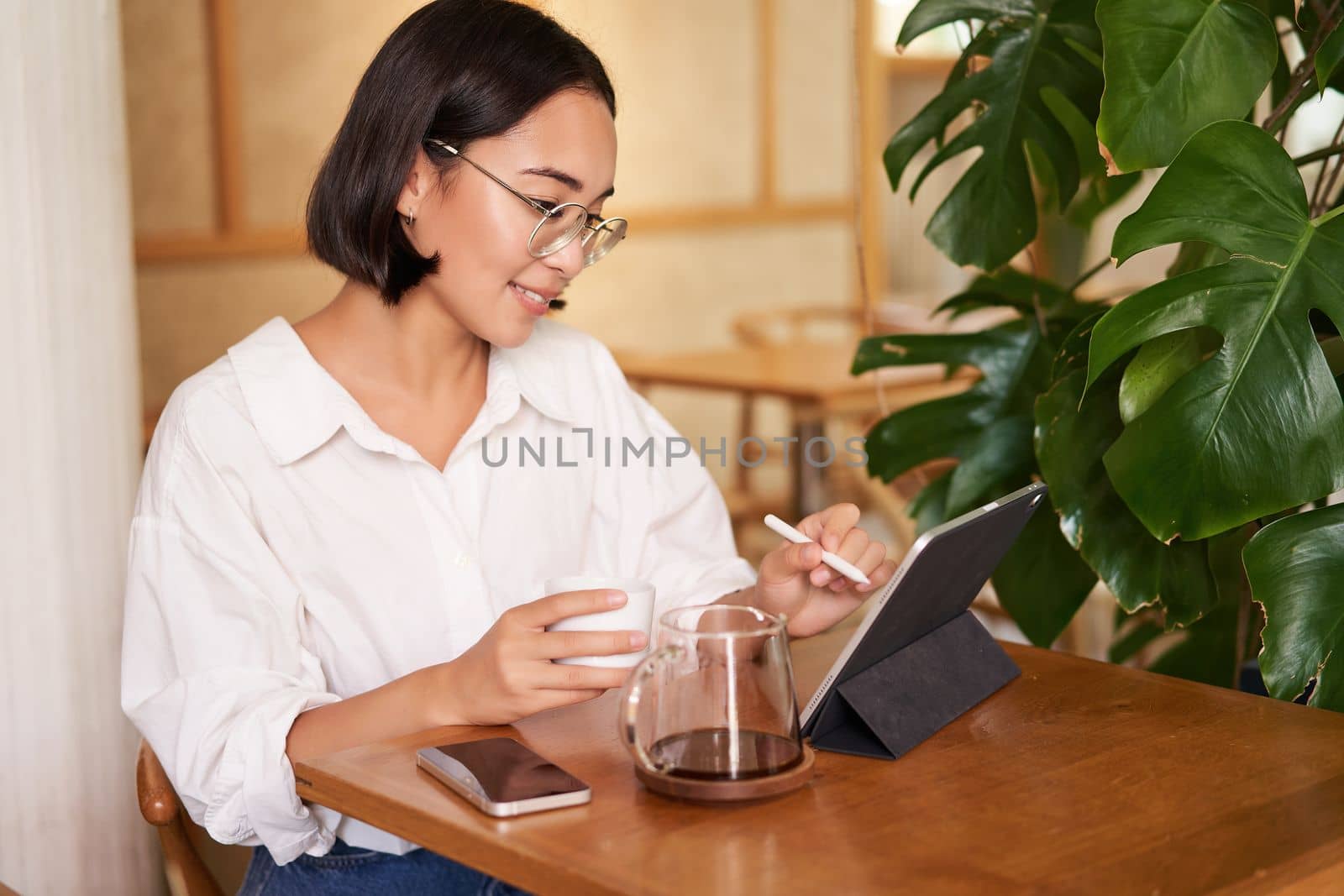 Asian woman in glasses, watching smth on digital tablet, drinking coffee in a cafe and smiling, working remotely.