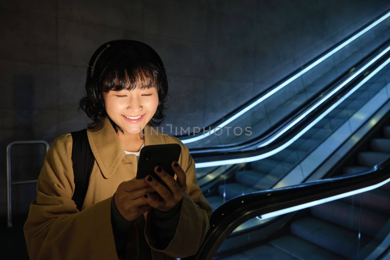 Happy asian woman in subway, standing near escalator, holding smartphone and listening music in headphones, smiling pleased.