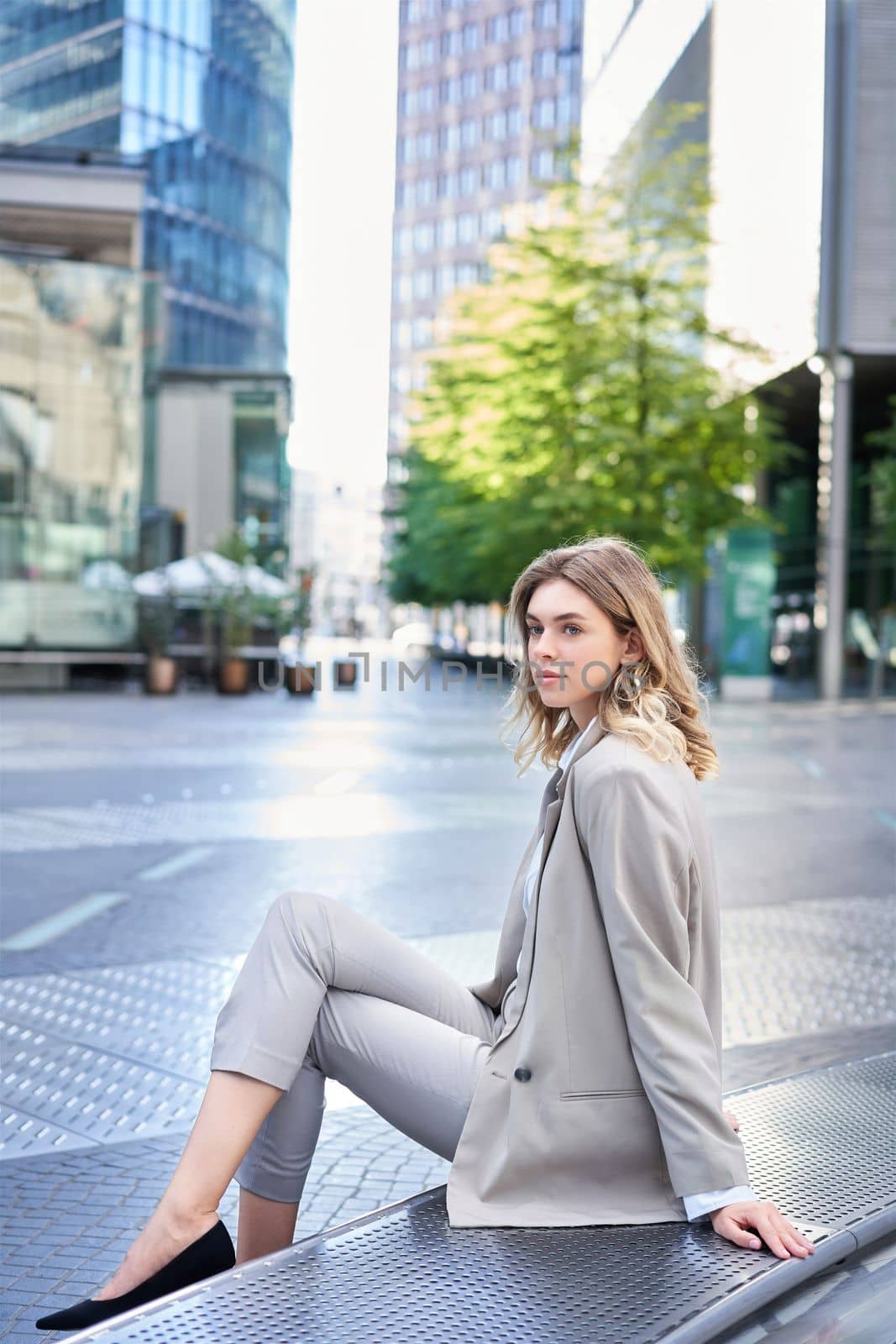 Vertical shot of successful young lady in suit, sitting outdoors, waiting for someone outside office. Employee relaxing on fresh air.