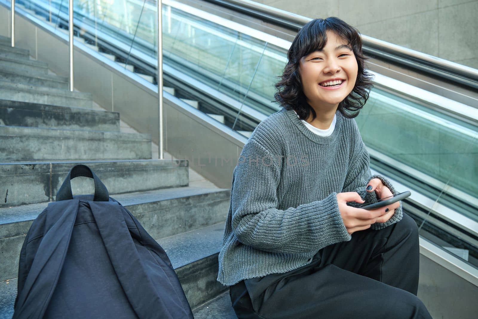 Stylish young girl student, sits on stairs with smartphone and backpack, laughs and smiles, texts message, uses social media.