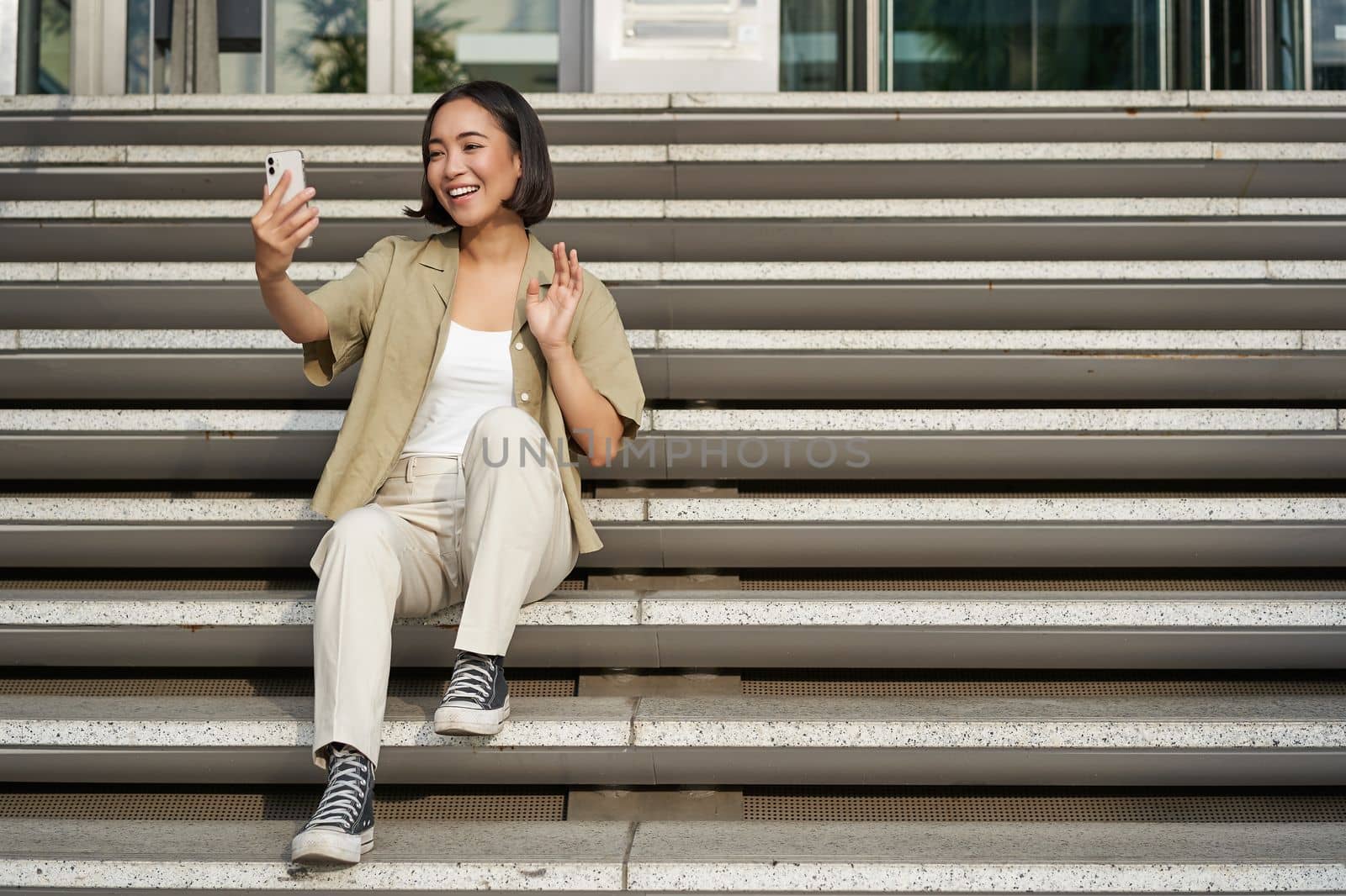 Portrait of asian beautiful girl takes selfie on smartphone. Young korean woman sits on stairs outdoors and makes photos on mobile phone.